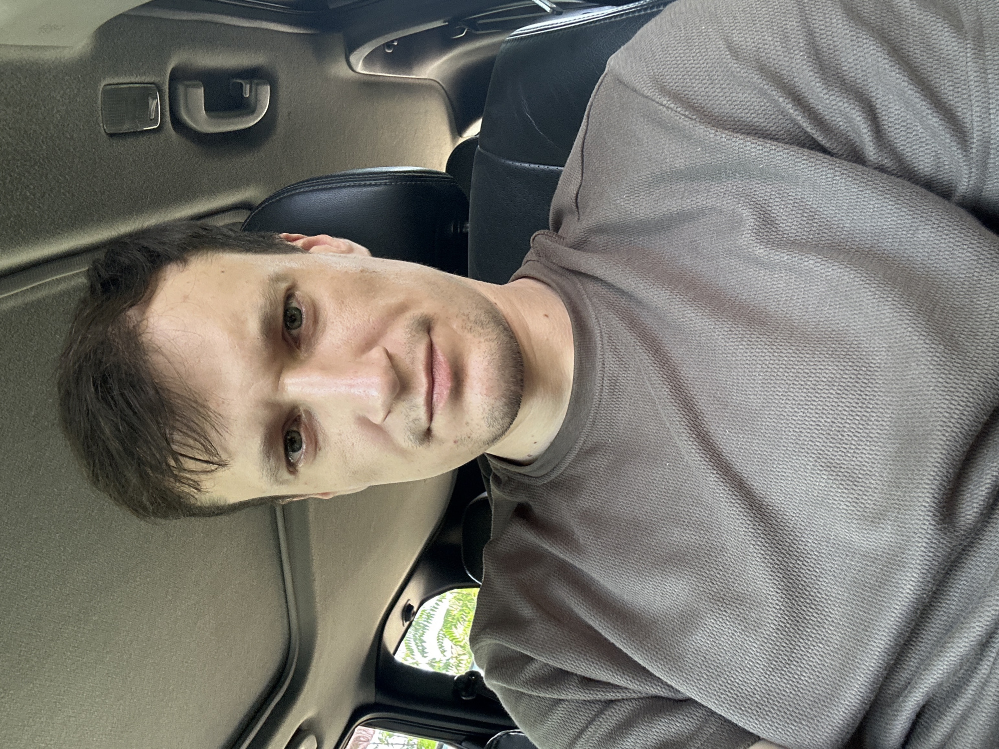The image shows a young adult male with short brown hair and light-colored eyes, sitting in the driver's seat of a vehicle. He is wearing a brown, textured t-shirt. The interior of the car is visible, including the headliner with a sunroof, a handle on the roof, and a black leather seat. Outside the car, through the window, foliage and trees can be seen. The scene appears to be in a natural setting, possibly on a sunny day. There is no specific indication of the location being in Makiv, Ukraine.