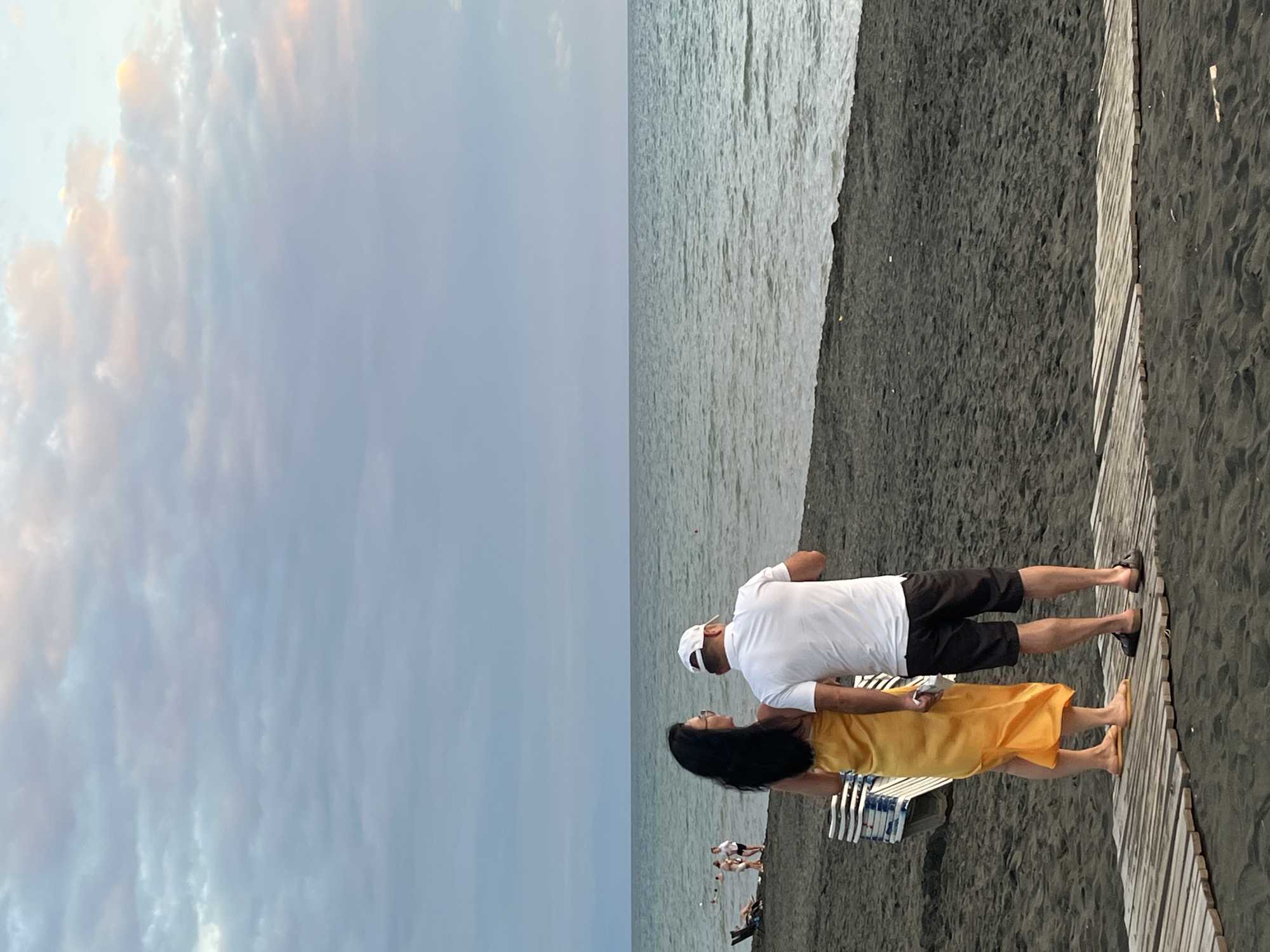 The image depicts a couple standing on a dark sand beach in Ureki, Georgia. The man, wearing a white t-shirt and black shorts, has a white cap on his head. The woman, beside him, is wearing a flowing yellow dress. They are facing away from the camera, looking out at the calm sea. The sky above is overcast with clouds, some of which are tinged with pink and orange from the setting sun. In the background, there are a few other people on the beach, and a stack of lounge chairs. A wooden walkway leads across the sand in the foreground. The overall scene is serene and captures a moment of quiet contemplation by the Black Sea.