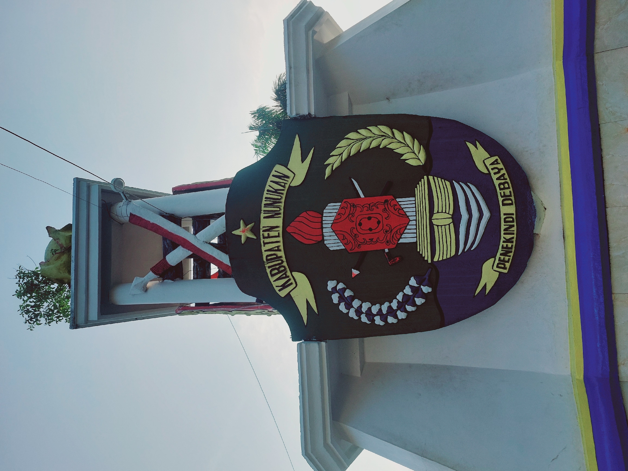 The image shows a close-up, low-angle view of a large, colorful emblem mounted on a white structure, likely a monument or building in Nunukan, Indonesia. The emblem is predominantly black with yellow, red, blue, and white elements. At the top of the emblem, a yellow banner reads "KABUPATEN NUNUKAN" in black lettering, with a red star above it. The central part of the emblem features a torch with red flames, a red shield with intricate designs, and crossed swords behind it. To the left of the shield is a white and purple laurel wreath, and to the right is a yellow laurel wreath. At the bottom of the emblem, another yellow banner reads "PENEKINDI DEBAYA" in black lettering. The white structure supporting the emblem has white columns and red and white striped accents. In the background, the sky is a pale blue, and a few green leaves and branches are visible at the top of the structure, suggesting it is outdoors. The lower part of the image shows a base with yellow and blue stripes. The overall scene conveys a sense of civic pride and local identity, as the emblem likely represents the regency of Nunukan.