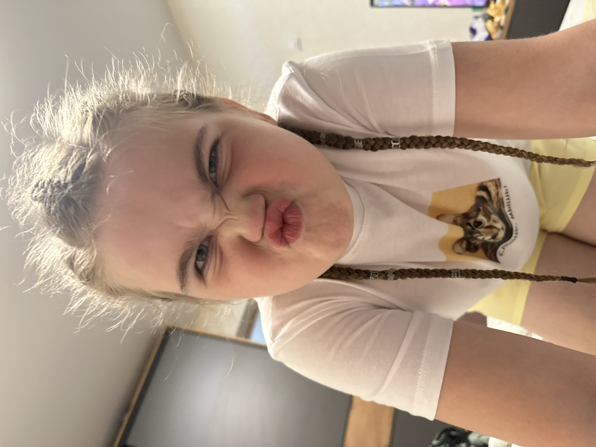 The image features a young person, gender female, appearing to be a child or pre-adolescent, looking directly at the camera from a low angle perspective. She has light-colored hair, styled in two visible braids that fall forward over her shoulders, adorned with several small, metallic, cylindrical beads. Numerous wispy flyaway hairs are visible around the crown of her head, brightly illuminated. Her eyes are light-colored, and she is making an exaggerated, playful expression: her lips are tightly puckered into a small "O" shape, her nose is crinkled, and her eyes are squinted and angled downwards.
She is wearing a plain white short-sleeved t-shirt. On the front of the shirt, a graphic depicts a cat's face (possibly a kitten) in shades of brown and white. Below the cat graphic, partially obscured text appears to be written in Cyrillic script. She is also wearing yellow shorts.
The setting is an indoor residential space. The background on the left side of the image consists of a dark, vertical surface, possibly a wardrobe or door, with a lighter section above it. On the right side, a plain, light-colored wall is visible. In the far right background, a blurred television screen displays colorful light, and some indistinct objects are present on a surface below it. The ceiling above the subject is plain and light-colored. The lighting appears to be ambient indoor light, with notable highlights on the subject's hair. There are no visual elements within the image (such as landmarks, unique architecture, or specific signage) that allow for the identification of the city of Chaikovsky, Russia, or any specific geographic location.
