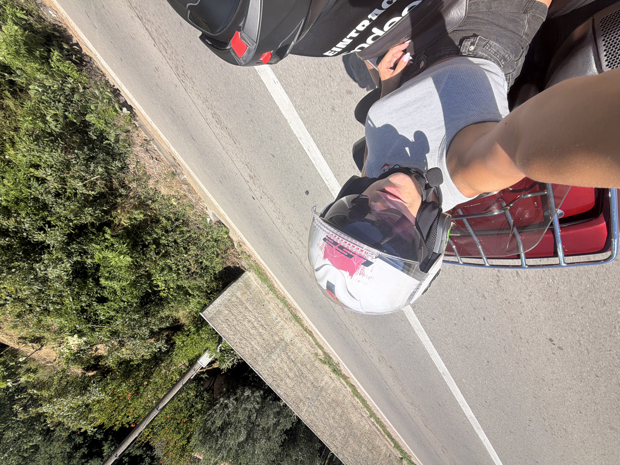 An overhead shot captures a person riding a scooter along a paved road in Mišići, Montenegro. The rider, wearing a white helmet with pink and black accents and sunglasses, is seen from the chest up. They are wearing a light gray t-shirt and black shorts. Sunlight casts a shadow on the rider and the road. To the right of the rider, another scooter helmet is visible. The environment consists of a road bordered by lush green vegetation and a low, textured stone wall on the left. The sky is a clear blue.