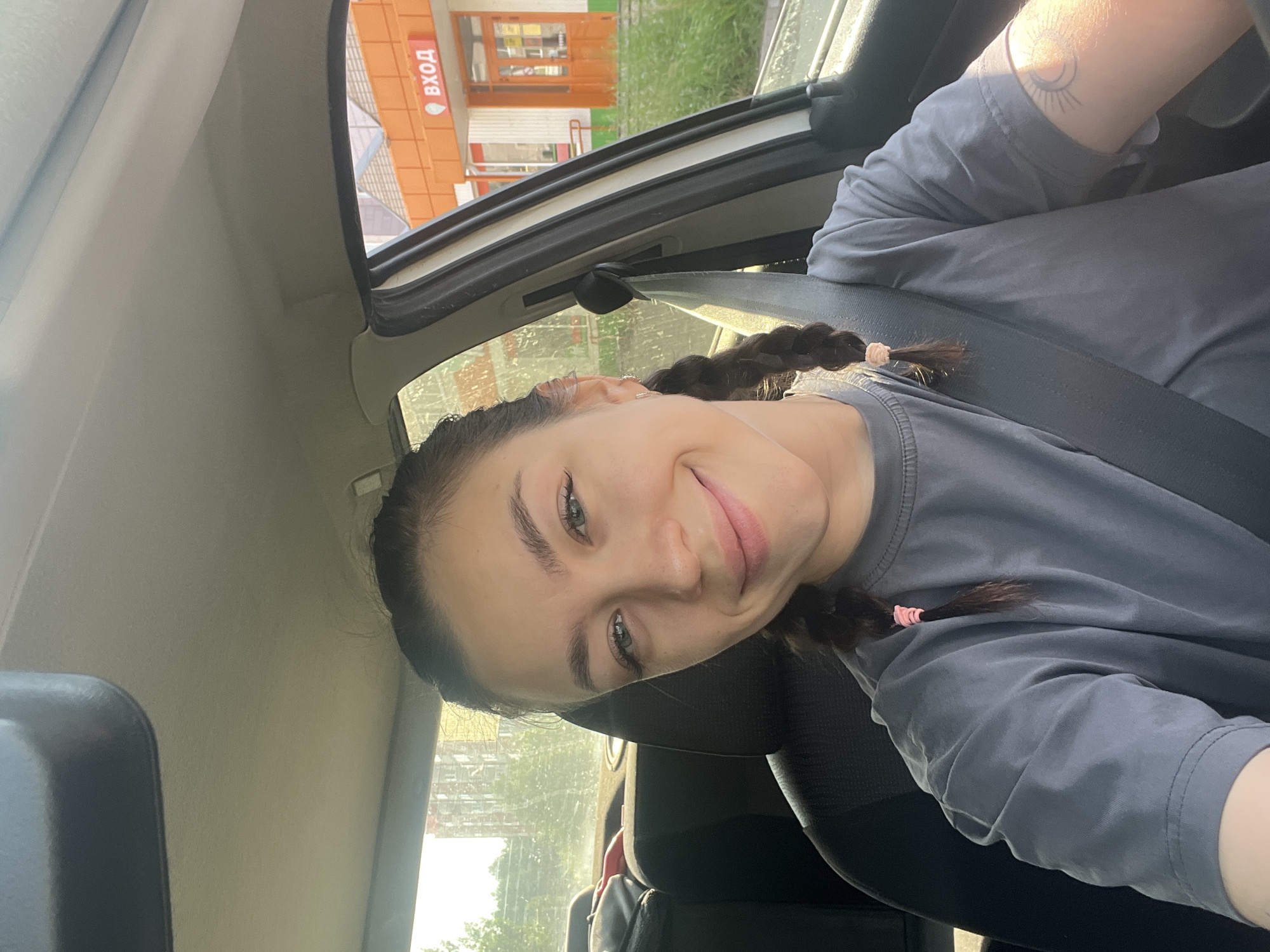 A young woman with braided brown hair, wearing a grey t-shirt and a seatbelt, is taking a selfie inside a car. She has green eyes and is smiling. In the background, through the car window, a street scene is visible with a building with an orange facade and a sign that reads "ВХОД" (Entrance) in Russian. Trees and a tall, modern building are also in the distance, suggesting an urban environment. The scene appears to be in Chaikovsky, Russia.
