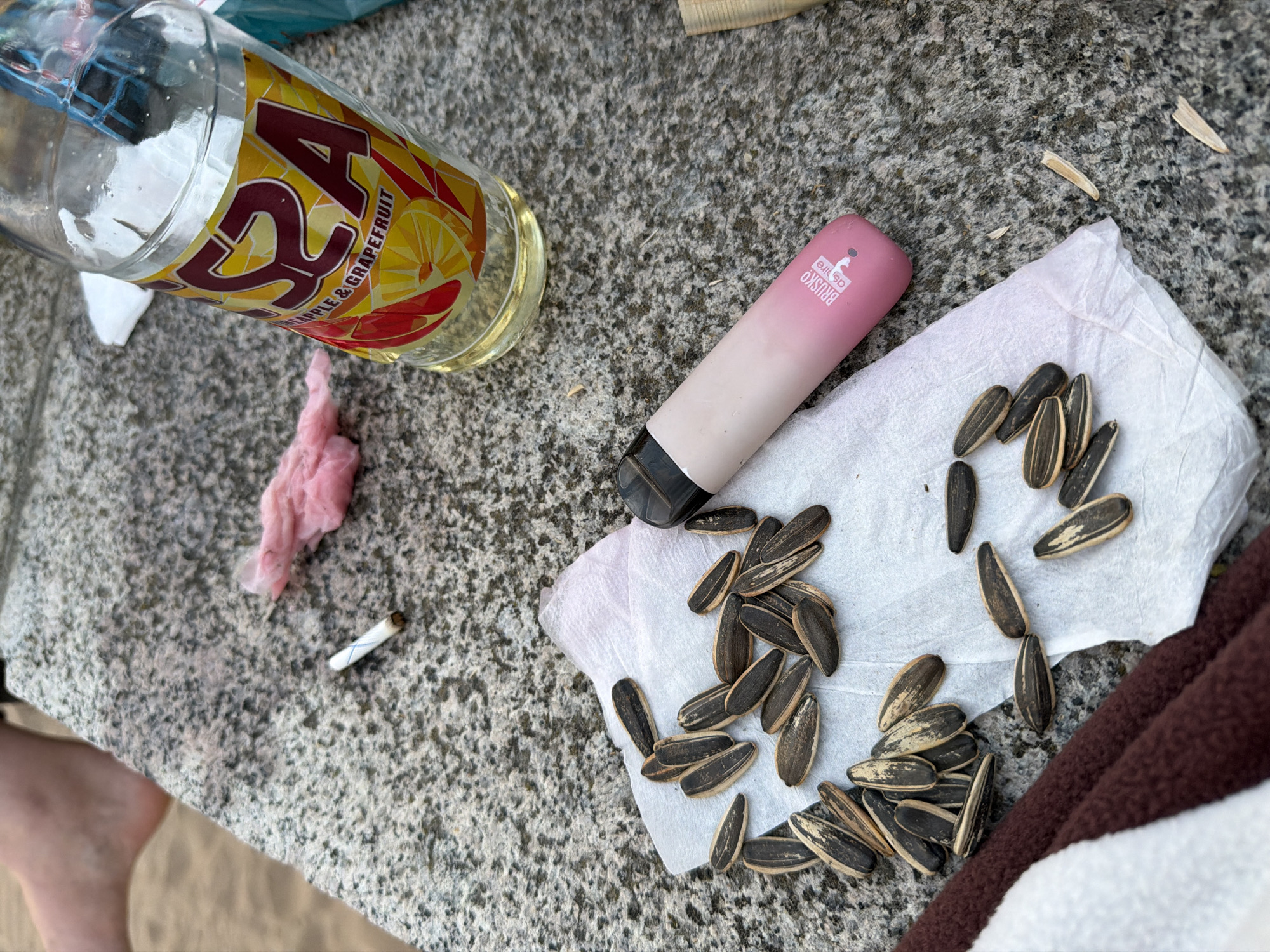 The image captures an overhead view of various personal items scattered on a textured, light gray, possibly granite or concrete surface, indicative of an outdoor setting in Saint Petersburg, Russia.
On the left, a clear glass bottle, approximately half-filled with a translucent yellow liquid, lies horizontally. Its label is prominently yellow and red with a mosaic-like pattern, displaying large red text that appears to read "2SA" and smaller text below stating "PINEAPPLE & GRAPEFRUIT."
To the right, a pink and white disposable vape pen is visible, positioned diagonally. Its side features a white logo and text that reads "BRUSKO as life." Beneath the vape pen, a white paper towel or napkin holds a pile of black and white striped sunflower seeds. Several individual sunflower seeds and empty shells are scattered both on and around the napkin, as well as on the gray surface, suggesting recent consumption.
Further to the left, near the bottle, a small, crumpled pink item, possibly a piece of fabric or tissue, rests on the surface. Below this, a discarded cigarette butt with a distinct blue cross pattern on its filter is present.
In the bottom left corner, a portion of a person's bare foot or ankle is visible, implying human presence. The bottom right corner shows a segment of a brown, textured material, likely a blanket or towel.
The bright, even illumination suggests the scene is set during daylight hours. The arrangement of items indicates a casual, relaxed outdoor activity, such as a picnic or gathering, with a focus on leisure and snacking.