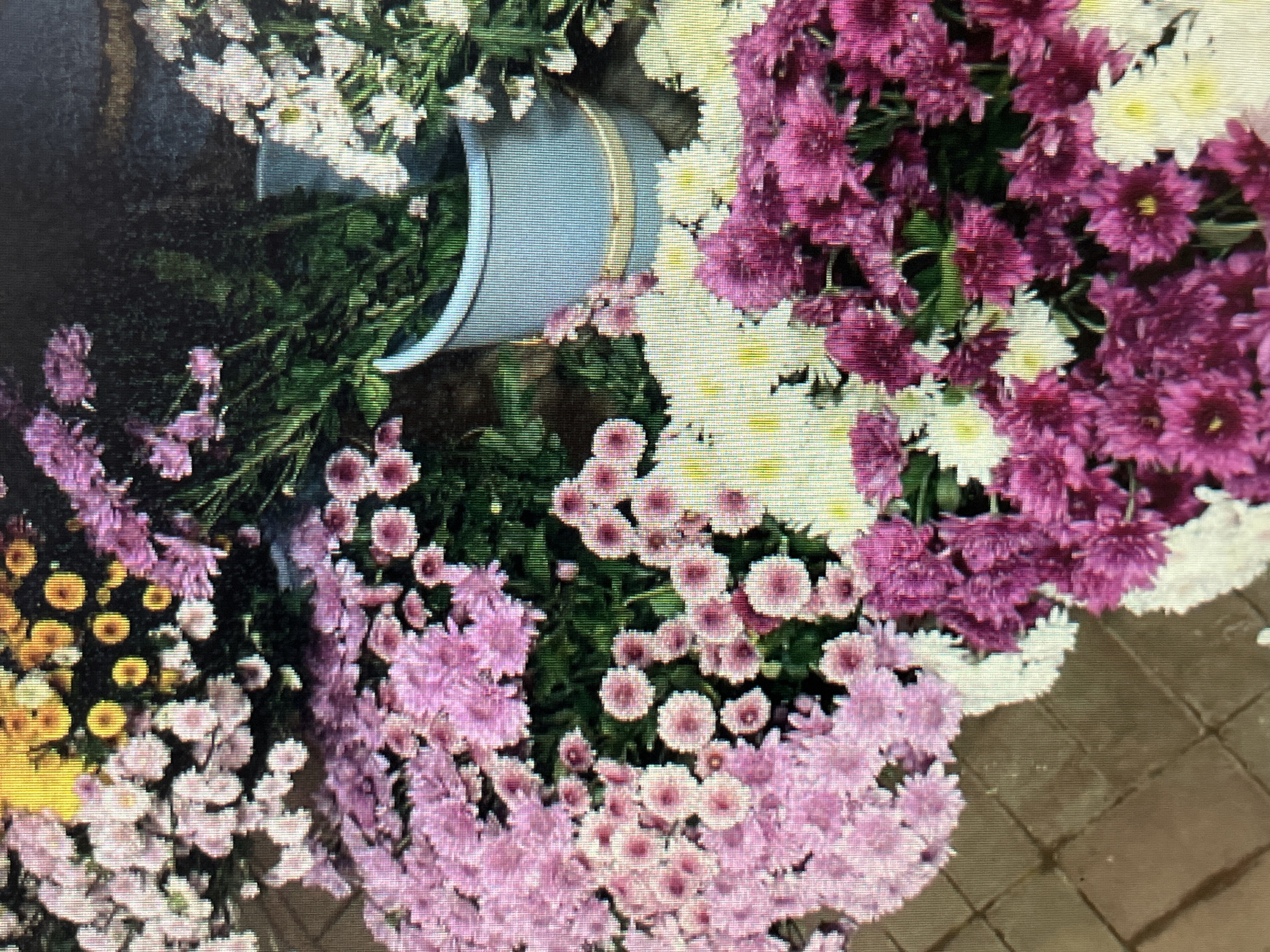 A close-up, overhead shot shows a vibrant display of chrysanthemums in various shades of pink, yellow, and white. The flowers are densely packed, with some stems visible. In the background, a light blue bucket, possibly containing water, holds more greenery and white flowers. The ground beneath the flowers appears to be paved with square, brown tiles. The scene is likely a flower market or shop, but the specific location in Sejahtera, Indonesia, cannot be determined from the image alone. There are no people or discernible actions in the frame.