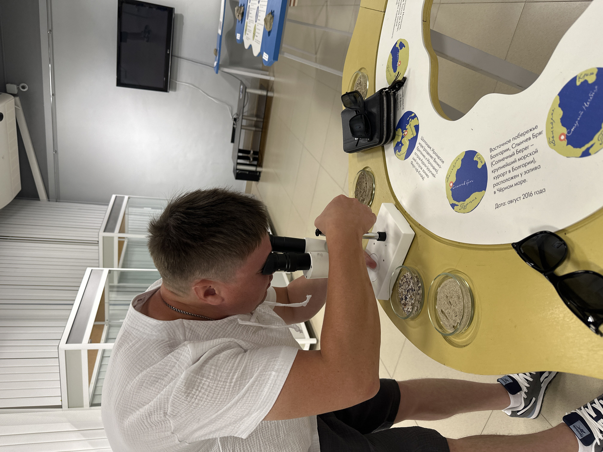 In the city of Chaikovsky, Russia, a man is intently examining sand and shell fragments under a microscope. He is wearing a white, textured short-sleeved shirt and dark shorts, with gray and white athletic shoes. The microscope is positioned on a yellow, curved table, which features several petri dishes containing the samples. Next to the microscope, a black wallet with sunglasses on top rests on the table. The table is also decorated with circular insets, each displaying a map of a body of water with text in Russian. The background reveals a bright room with white blinds on the left and a grey wall with a television screen on the right. This scene appears to be within a museum or educational exhibit, possibly related to natural sciences or local geography, given the maps and samples. The overall atmosphere is one of focused observation and learning.