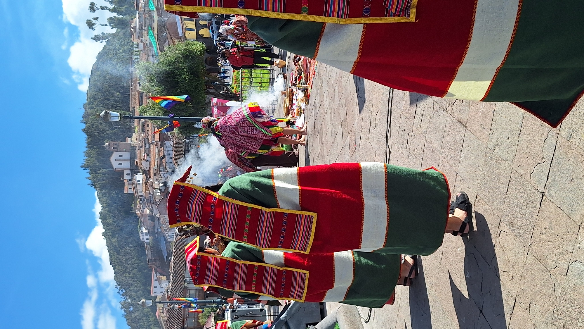 Here is a description of the image:
The image shows a scene in a city, likely in Peru. A group of people is performing a ceremony involving a fire, with smoke rising in the background. The performers are wearing traditional garments, including vibrant red, green, and white colored fabrics. One individual is wearing a pink patterned textile. Some are barefoot. The setting includes a paved area with buildings and a hillside visible in the distance. A rainbow flag and lampposts are also visible.