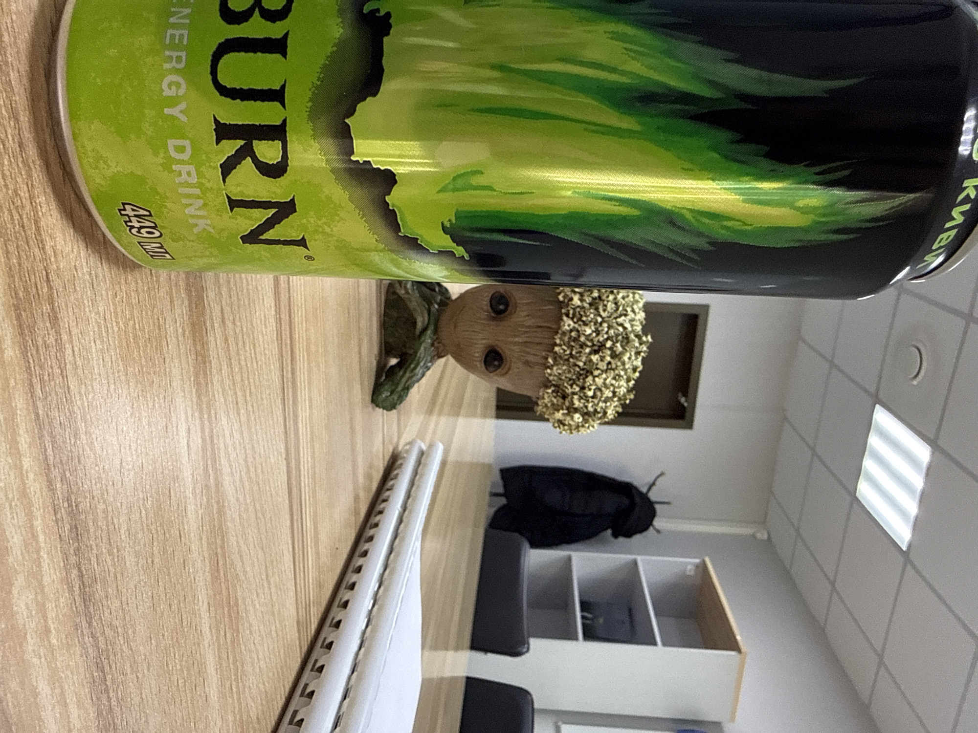 The image shows a can of BURN energy drink lying on a light brown wooden surface. Beside the can is a small Groot figurine. In the background, there is a white cabinet with compartments, a black coat hanging on a hook, a black office chair, and a tiled wall with a window. The location is Krasnoyarsk, Russia, based on the Cyrillic letters on the can.
