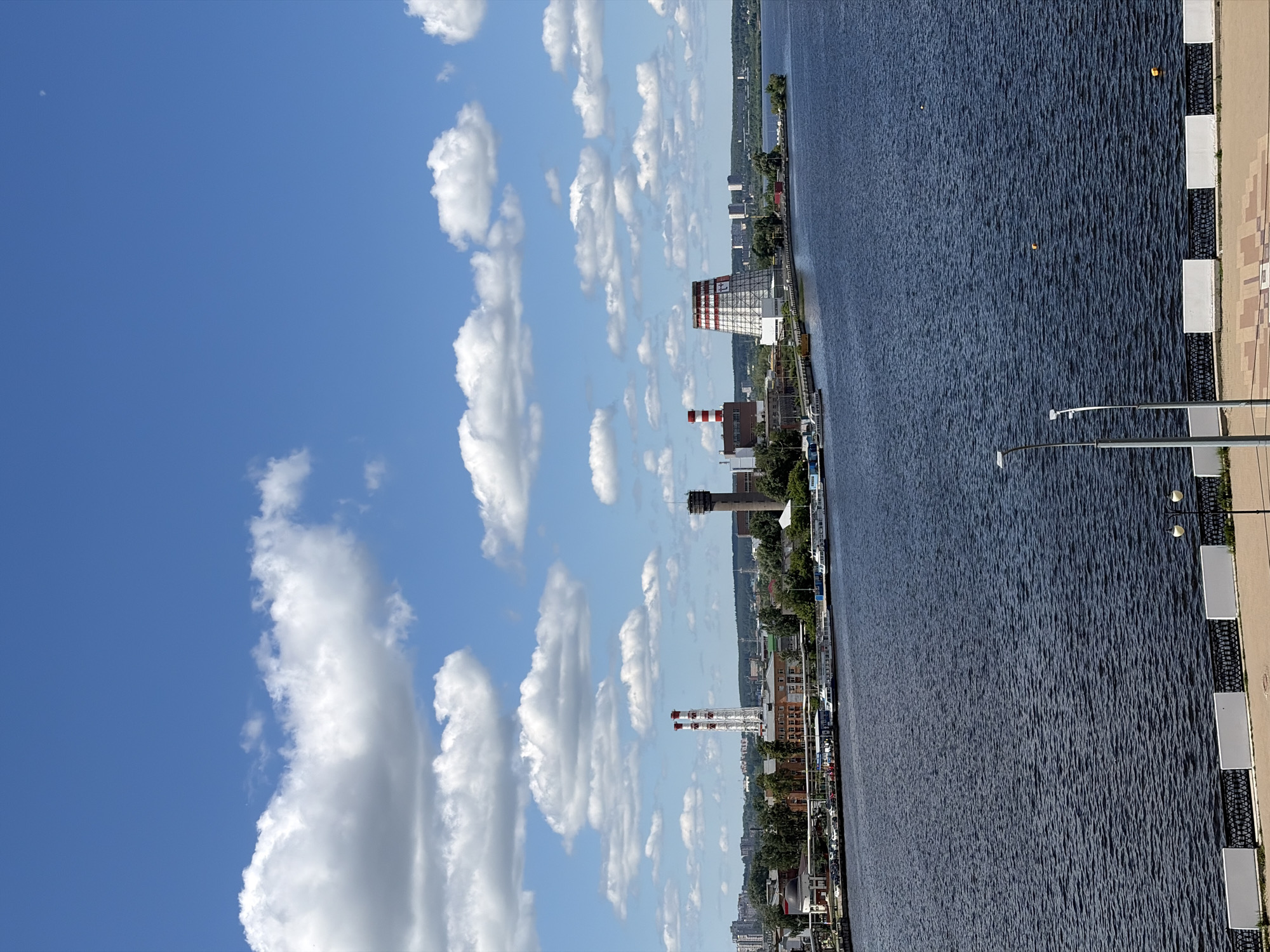 The image displays a panoramic view of a city waterfront, identified as Izhevsk, Russia, under a bright blue sky filled with scattered white cumulus clouds. A wide expanse of dark blue water dominates the midground, its surface showing ripples, with a few small yellow buoys floating within it.
In the foreground, along the right bank of the water, a light-colored, patterned paved walkway is visible. This walkway is bordered by a dark metal fence featuring regularly spaced white rectangular panels. Two streetlights with double light fixtures stand along this promenade.
The far bank, to the left across the water, showcases the urban landscape of Izhevsk. A mix of industrial and commercial buildings lines the shore, including a large white building with distinctive red striped accents near its roof. Several prominent smokestacks rise from industrial complexes; one is notably tall with red and white stripes, while another is a reddish-brown. A slender brown tower also stands out among the structures. The urban scene is interspersed with green foliage from trees and shrubs, and a road with parked vehicles is discernible along the bank. In the background, more distant buildings and forested hills complete the horizon.
The scene captures a clear, sunny daytime view with no specific human activity or visible text. The overall atmosphere appears calm and bright.