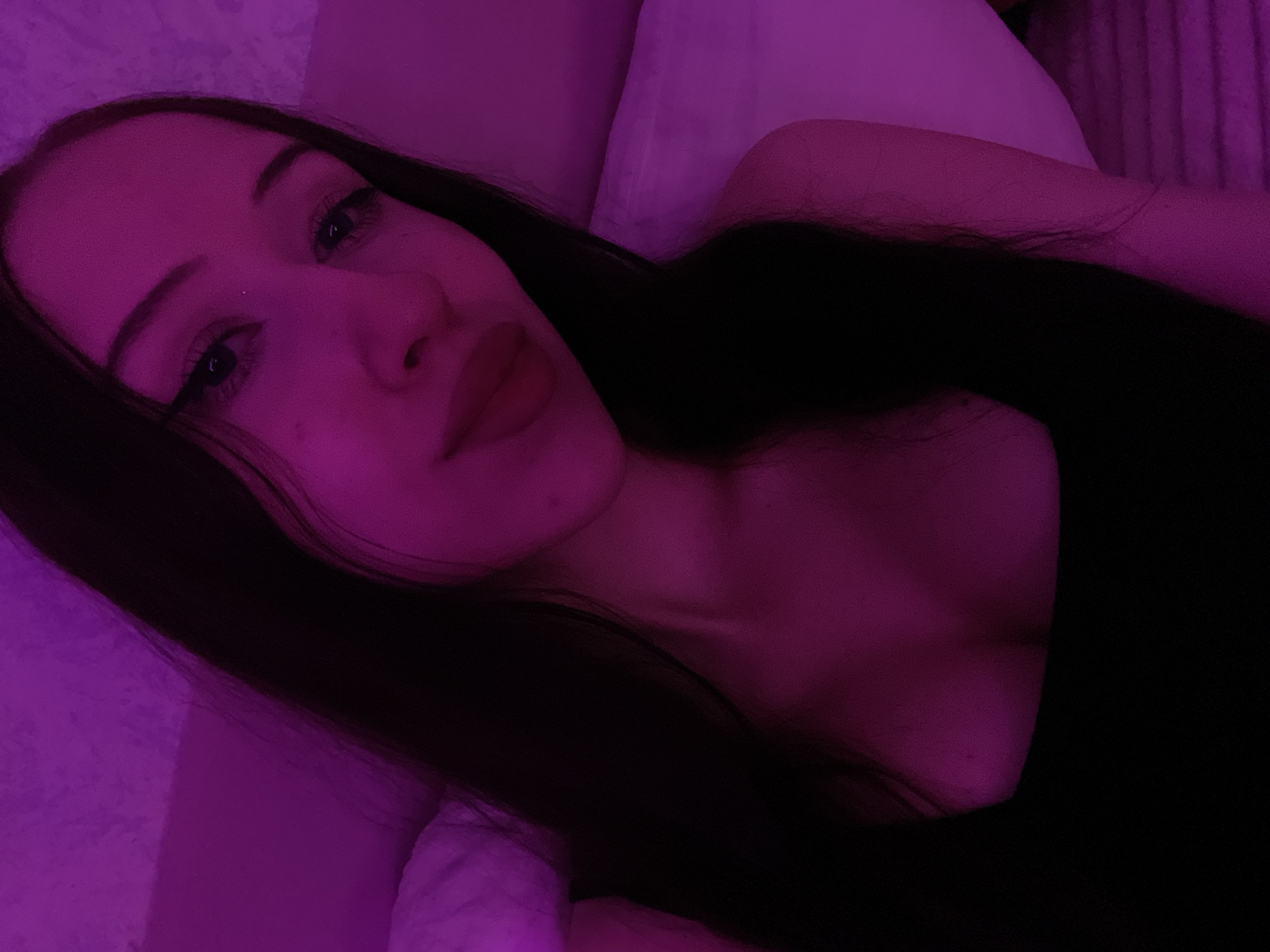 The image is a close-up selfie of a young woman with long, dark hair, taken indoors under purple lighting. She is wearing a black top. Her face is partially visible, and she has on dark eyeliner and lipstick. The background appears to be a bed with pillows, and there is a textured wall behind her. The setting is domestic. The image does not provide any information about the city of Votkinsk, Russia.