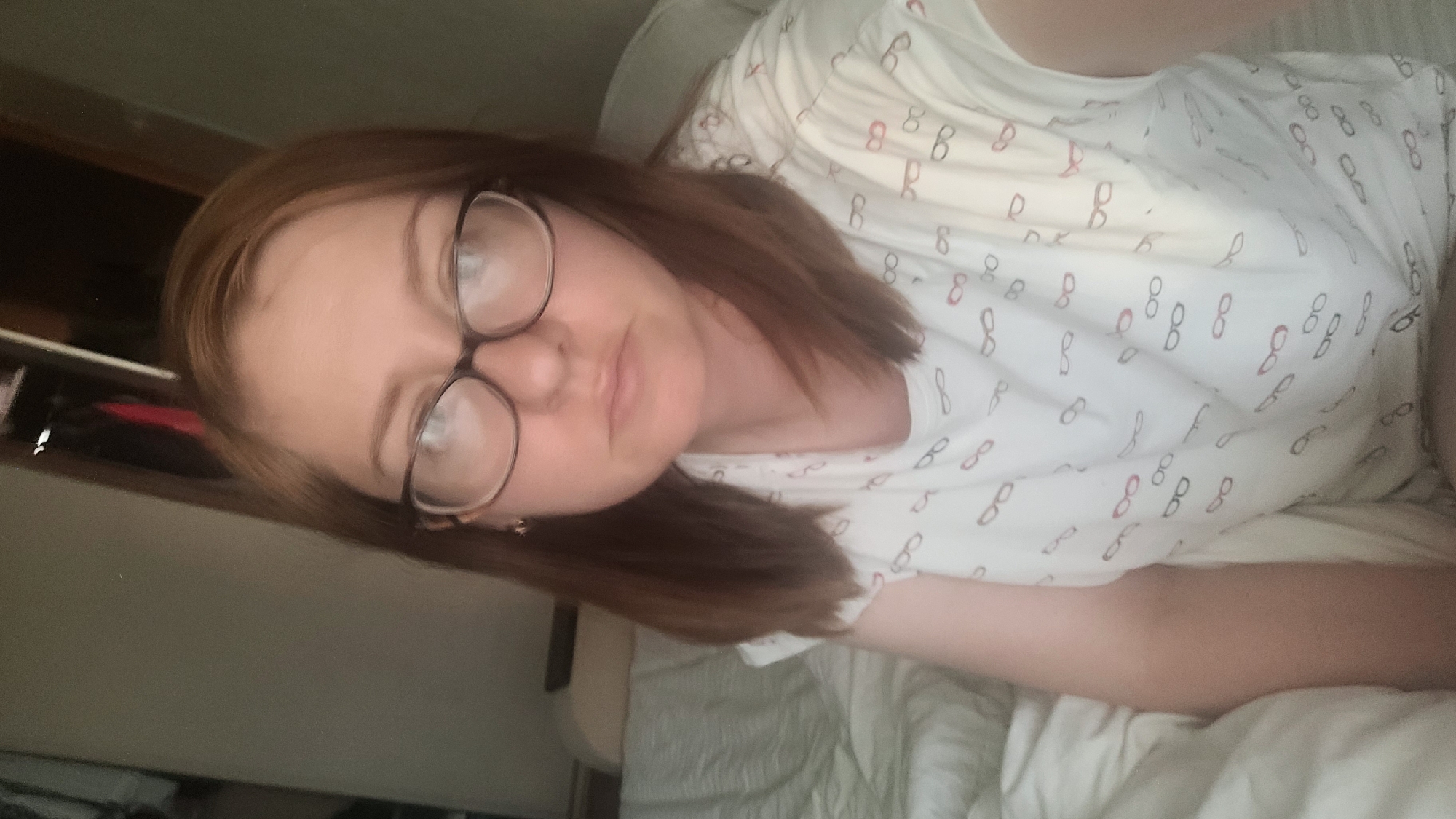 This media file is a portrait-style, sideways selfie of a person wearing glasses and a white t-shirt with a pattern of glasses printed on it. The subject has light skin and shoulder-length brown hair. They are lying down, possibly in a bed, as indicated by the white bedding visible in the lower portion of the image. The lighting suggests it might be daytime, but the indoor setting makes it difficult to determine the exact time of day or weather conditions. The overall mood appears relaxed and casual. The visible text in the image consists of the repeated glasses pattern on the t-shirt. The orientation of the image is rotated.