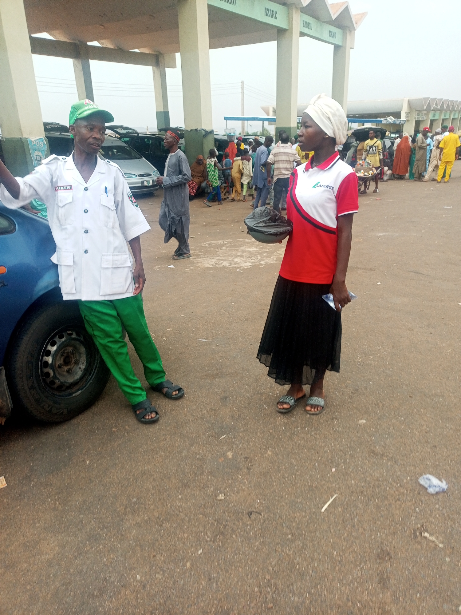 The image captures a scene in what appears to be a busy outdoor area, possibly a transit point, in the city of Gombe, Nigeria. Two individuals are prominently featured in the foreground.
A man dressed in a light-colored uniform with green trousers and a matching green cap is leaning against the left front tire of a blue car. A logo is visible on his uniform. He is wearing sandals.
To the right stands a woman with a white head covering, wearing a red and white shirt with "LAFARGE" printed on it, a black skirt, and sandals. She holds a dark-colored object in one hand and a small white object in the other.
In the background, a large group of people is gathered, with various clothing styles suggesting diverse backgrounds. Cars are parked and infrastructure is visible in the background, including overhead structures with what appears to be city names written on them. The ground is paved and shows signs of wear and some litter.