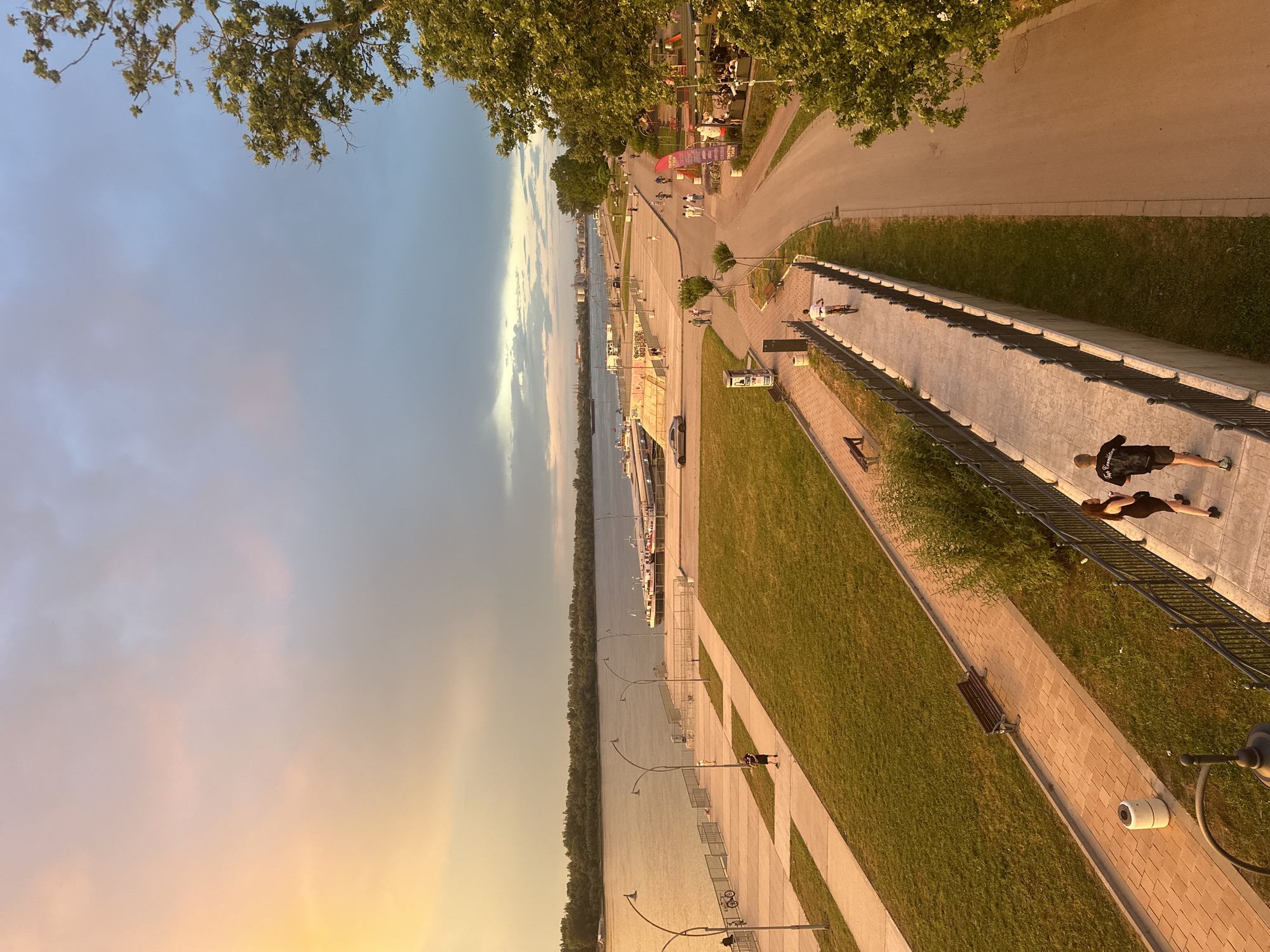 The image depicts a scenic view of the Ruse riverfront in Ruse, Bulgaria, during what appears to be late afternoon or early evening. The sky is a gradient of warm yellow and orange hues on the left, transitioning to cooler blues and grays on the right, with scattered clouds. A wide river, likely the Danube, dominates the left side of the frame, with a tree-lined embankment visible on the opposite bank. Several boats, including what appear to be barges and a passenger ship, are docked along the Ruse embankment.
In the foreground, a paved walkway runs parallel to the river, lined with lampposts and a metal railing. On this walkway, a man and a woman are jogging, with the man slightly ahead of the woman. Further along the walkway, a man is running, and further still, a person is riding a bicycle. On the grassy area between the walkway and the river, there are benches.
To the right of the walkway, a paved road curves through a park-like area. This area is populated with various people, some walking, some on bicycles, and others seated at tables in what looks like an outdoor seating area for a cafe or restaurant. Trees, some with dense green foliage, are scattered throughout this area and frame the right side of the image. The overall scene conveys a sense of leisure and activity along the riverfront.