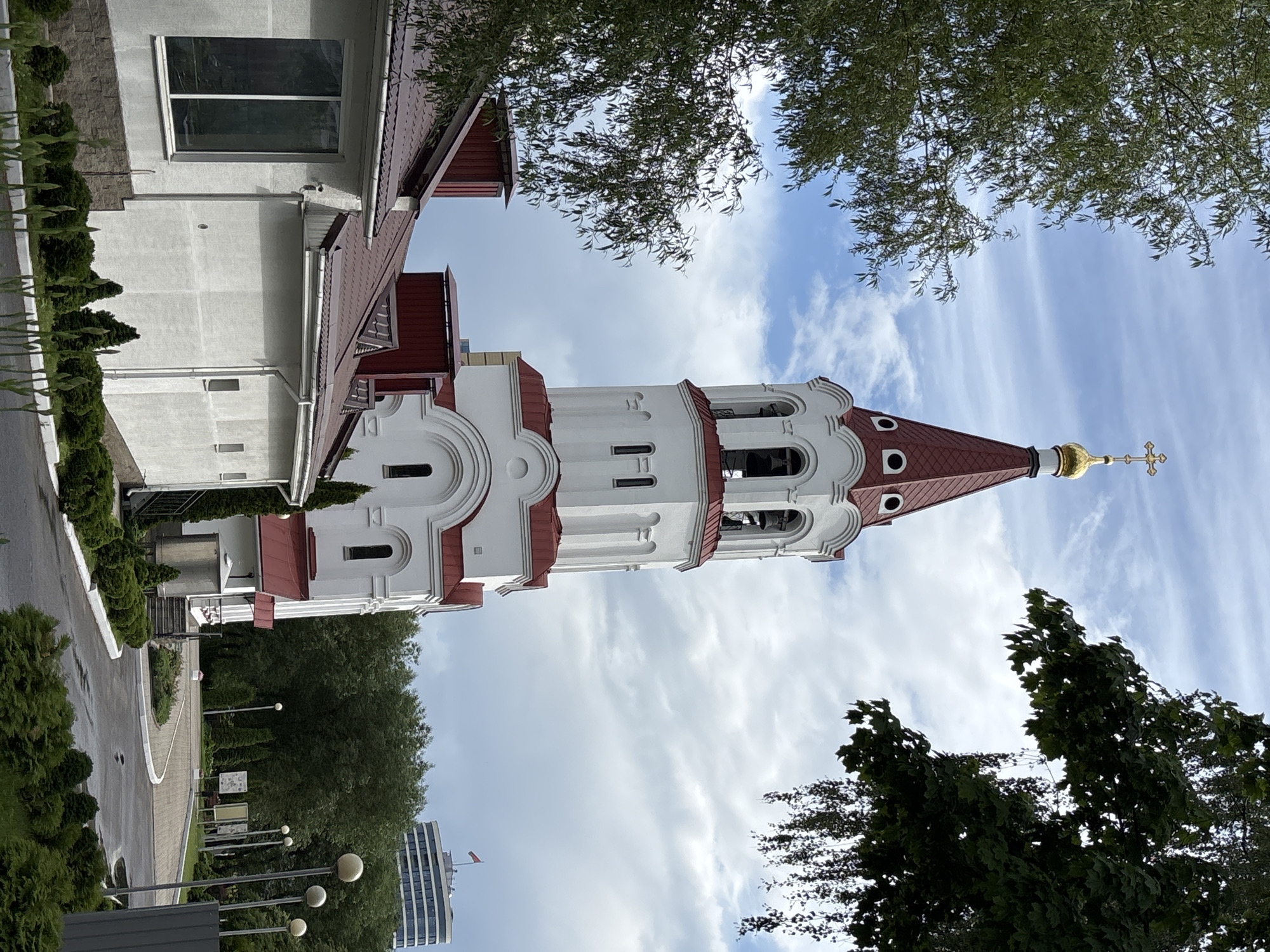 The image shows an upward-angled view of the Holy Spirit Cathedral in Minsk, Belarus. The cathedral, characterized by its white walls and red roofs, prominently features a tall bell tower topped with a golden dome and cross. The tower's architectural style includes arched openings, contributing to its distinctive appearance. A portion of an adjacent building with windows is visible on the left. The sky is partly cloudy with varying shades of blue. Tree branches frame the image, adding depth. In the distance, a modern building is visible behind a green area with street lamps. The scene appears to be taken during daylight hours.