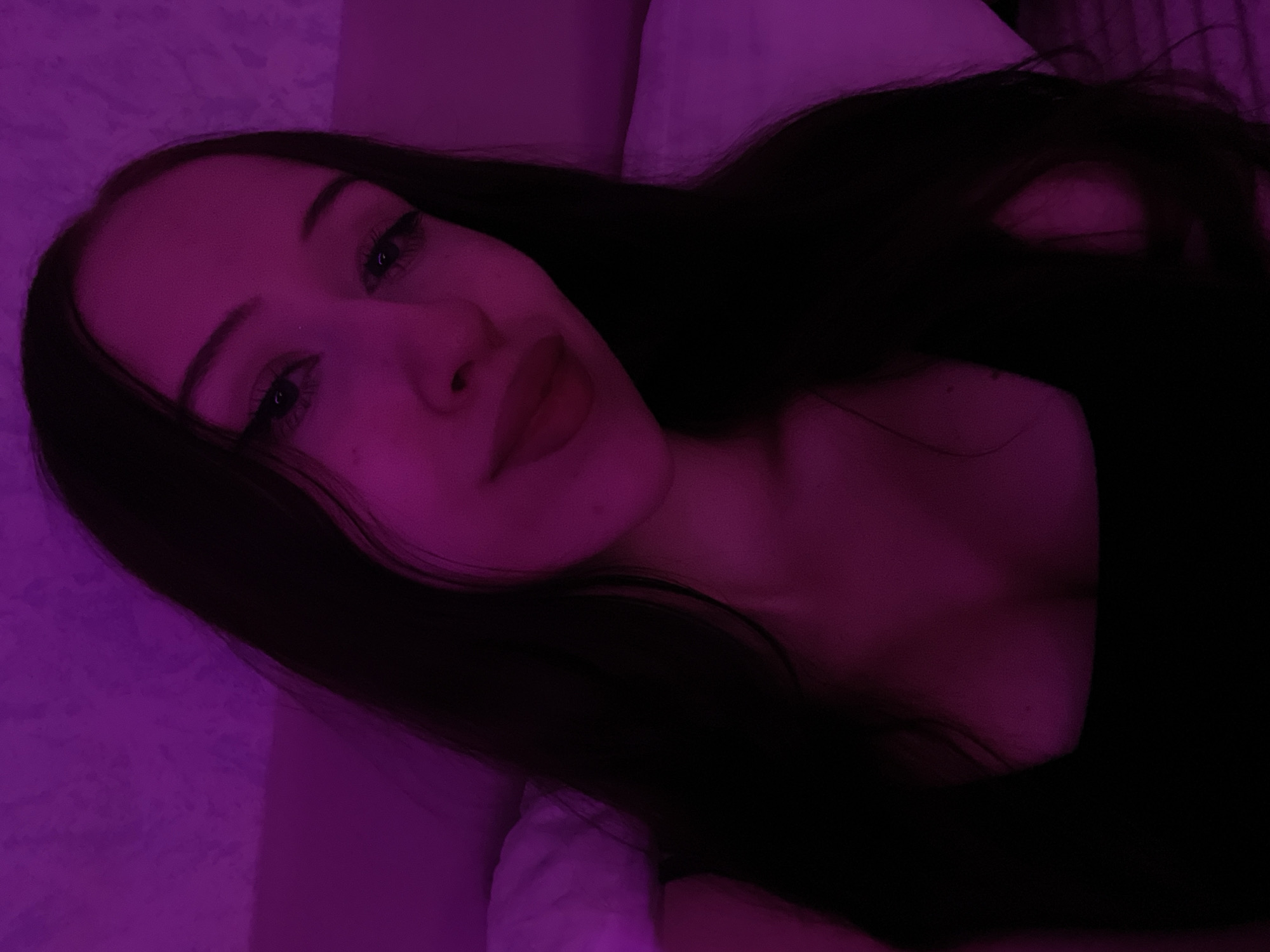 An indoor, close-up, selfie-style photograph shows a young woman with long, dark hair and fair skin. She is wearing a dark-colored top with a square neckline. The lighting in the image is predominantly purple, with some pink hues. The background appears to be a textured wall and a white surface, possibly a pillow or bed. The woman is looking directly at the camera. There is no discernible information in the image to suggest a location in Votkinsk, Russia.