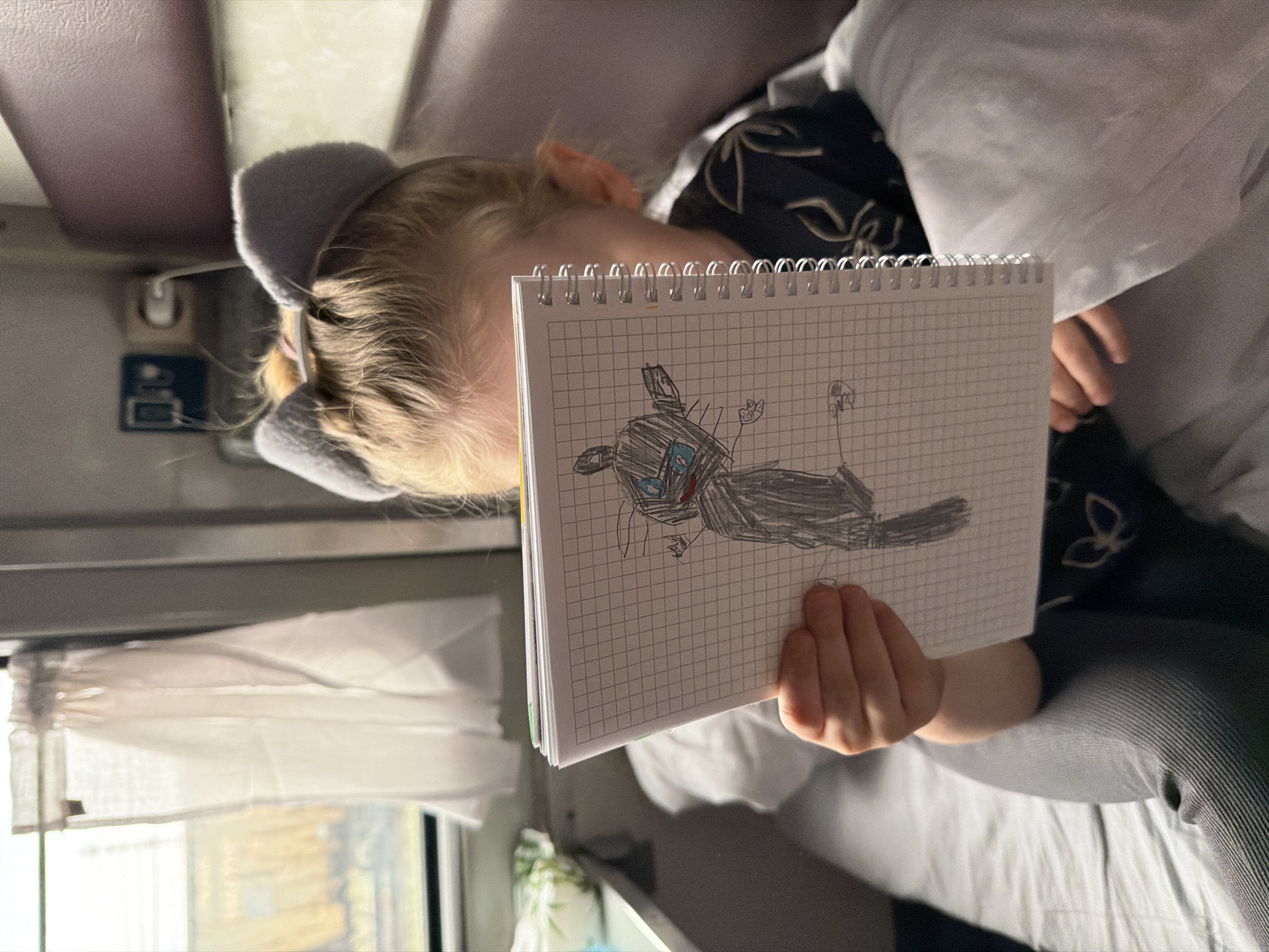 A young child, likely a girl with blonde hair, is captured indoors, holding a spiral-bound notebook. She wears a dark top with a light pattern and has a grey fluffy headband, possibly with "cat ears." Her face is largely obscured by the notebook, but her right hand holds it open, displaying a vibrant drawing on grid paper. The artwork depicts a dark gray, stylized creature or monster with distinct blue eyes and red facial markings, along with a tail.
The setting appears to be the interior of a vehicle, possibly a train, suggested by the design of the window with a sheer curtain on the left, and the overhead panels and a wall-mounted light fixture or button visible in the background. Natural daylight illuminates the scene from the window, indicating it is daytime. The child is partially covered by a light-colored blanket or clothing, and dark grey pants are visible. The image portrays a serene moment, possibly during a journey in Uzunovo, Russia, given the location context and typical mode of transport through such areas. No discernible text is visible in the image.