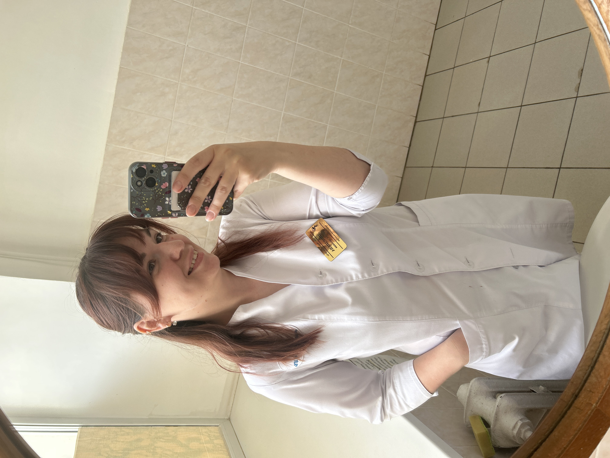A woman is taking a selfie in a bathroom. She is wearing a white coat with a name tag that reads "Электричка Васильевна Тюфякова," suggesting the location may be in Russia. The bathroom has beige tiles and a round mirror. A white radiator is visible near the sink. The woman is holding a phone with a floral-patterned case.