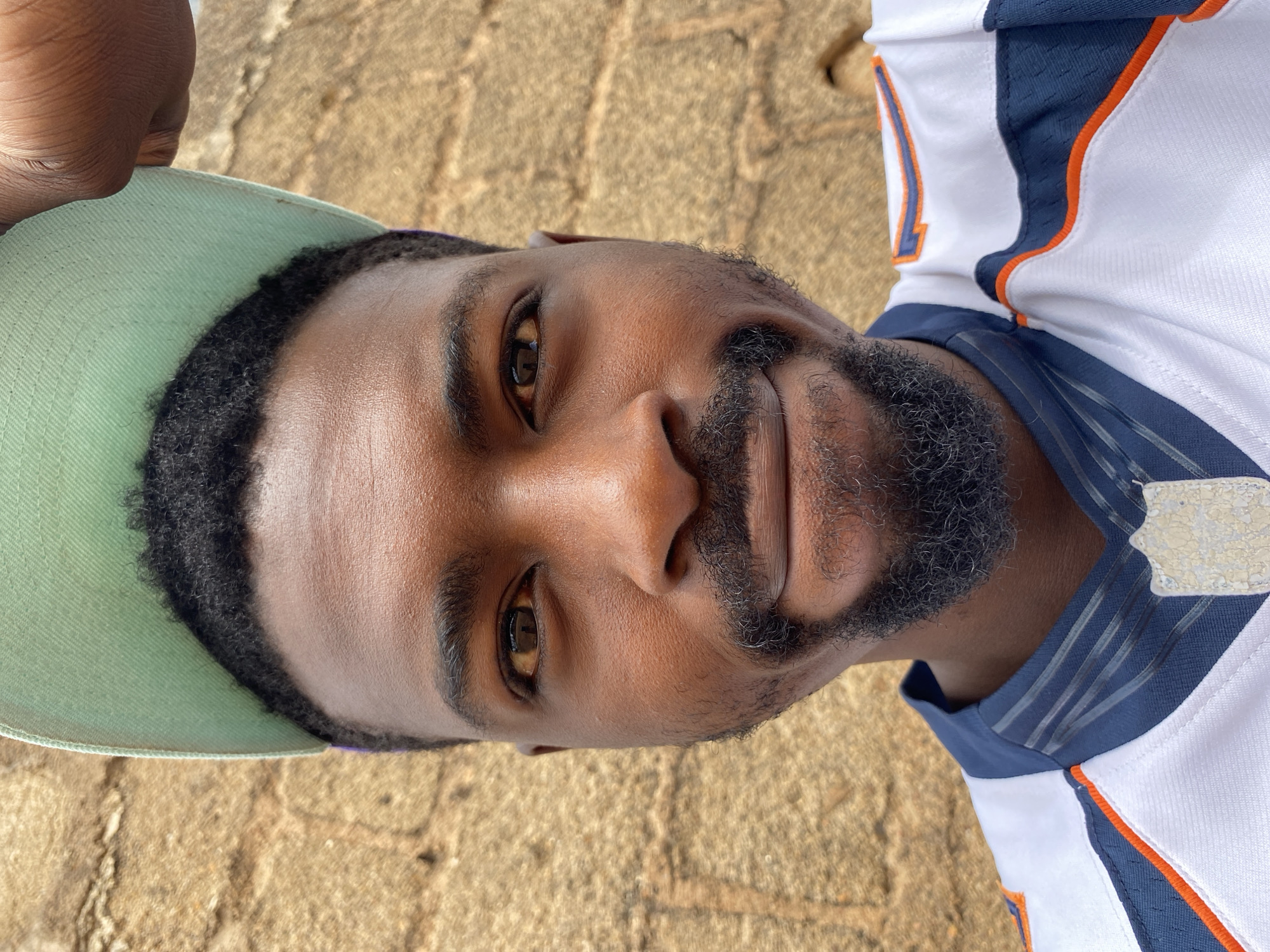 A close-up, eye-level portrait features a man with dark skin, looking directly at the camera with a neutral to slightly content expression. He is wearing a light mint green baseball cap and a sports jersey.
His facial features include dark brown eyes, a neatly trimmed beard, and a mustache. The jersey is predominantly white, with distinct blue side panels and orange trim. A partial number "1" is visible on the left side of his chest. A faded or worn patch, possibly a logo, is present on the right shoulder area of the jersey. The colors and style of the jersey evoke an American football uniform, potentially that of the Denver Broncos. A hand, matching the man's skin tone, is visible holding the brim of the cap on the left side of the frame.
The background consists of a textured, light brown or tan surface, possibly paving stones or ground, suggesting an outdoor setting. The bright, even lighting indicates daytime, possibly under open shade or an overcast sky, avoiding harsh shadows on his face. The scene is consistent with an outdoor environment in Jos, Nigeria.