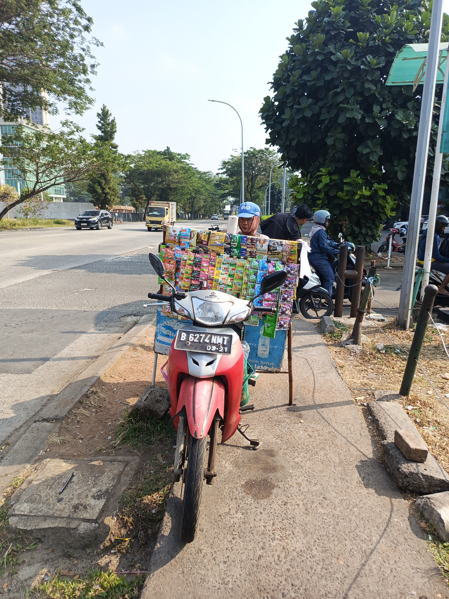 This is an eye-level, medium shot of a street vendor selling snacks from a modified motorcycle on a sidewalk in Tangerang, Indonesia. The vendor, a man wearing a blue cap, stands behind a rack of colorful snack packages displayed on his motorcycle. The motorcycle is red and white with the license plate "B 6274 NMT 03-21." Two people on motorcycles are visible in the background, one of them interacting with the vendor. The street is paved, with a concrete sidewalk and a grassy area. Trees and buildings line the street, with a blue sky overhead. The scene suggests a typical urban setting in Indonesia with local commerce taking place.