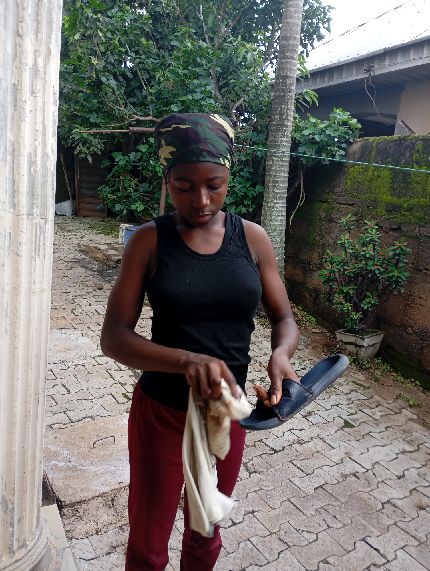 In the provided image, a young Black woman is depicted in an outdoor courtyard. She is wearing a black tank top, maroon pants, and a camouflage headscarf. The woman is holding a dark blue sandal in her right hand and a light-colored cloth in her left hand, appearing to be in the process of cleaning the sandal. The courtyard is paved with interlocking concrete blocks, and various elements of the environment are visible. To the left, a portion of a weathered, fluted column is present. Behind the woman, lush green trees and foliage dominate the background, with a hint of a building with a corrugated metal roof in the upper right corner. A stone wall covered in moss can be seen to the right, with a potted plant featuring small red flowers situated against it. The overall impression is of a residential or semi-rural setting. The image is a full-body shot of the woman, with the background providing context for her activity.
