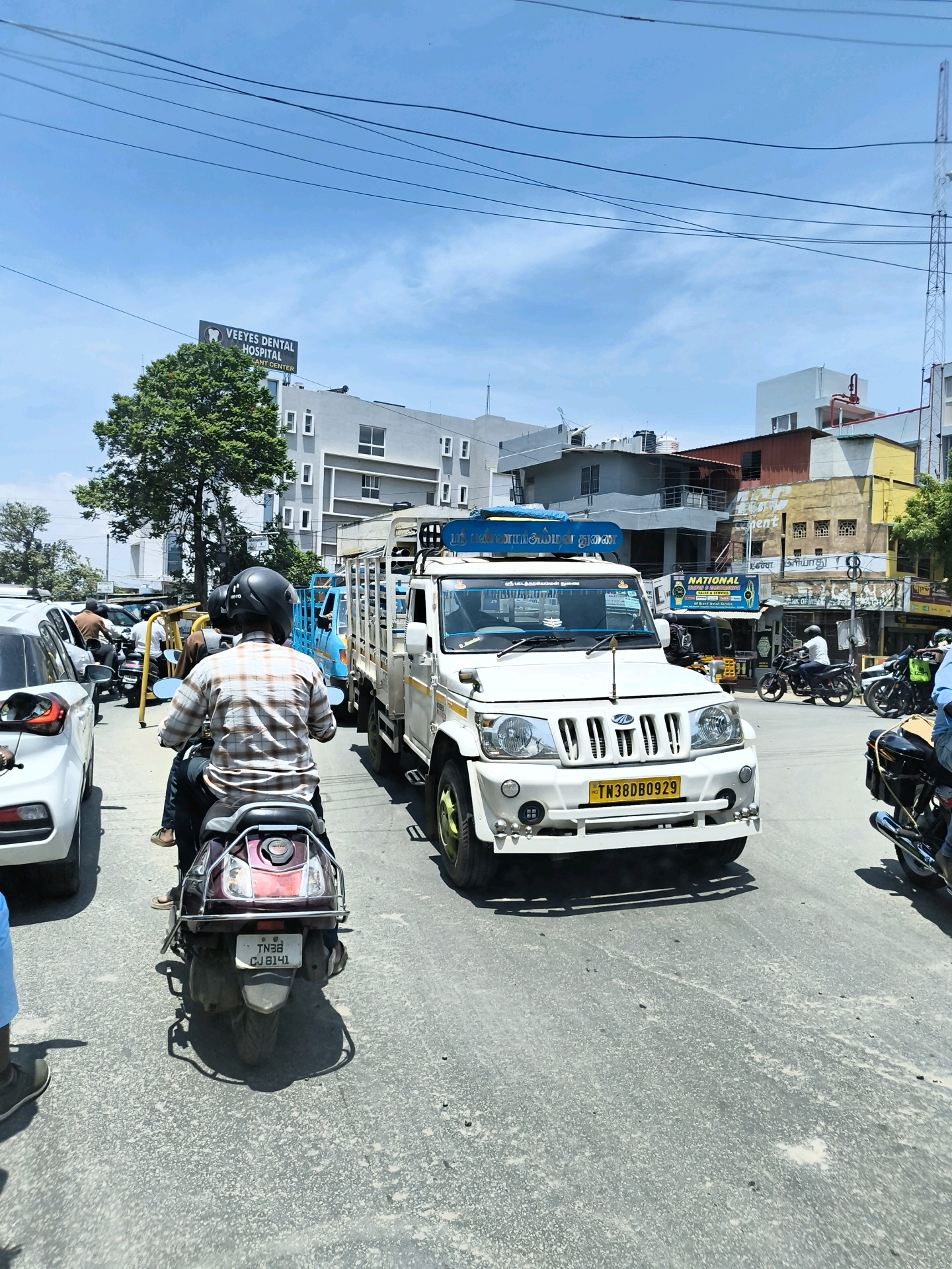 The image shows a traffic scene in Coimbatore, India. A white Mahindra Bolero pickup truck is in the foreground, with a license plate reading "TN38DB0929". The truck is in traffic on a paved road. Behind the truck, a person wearing a plaid shirt and a black helmet is riding a dark-colored scooter. Other vehicles, including a white car and a light blue truck, are visible in the background. Buildings line the street, including a structure with a sign that reads "VEEYES DENTAL HOSPITAL". The sky is blue with some clouds, and power lines are visible overhead. The overall scene depicts a busy urban street during the daytime.