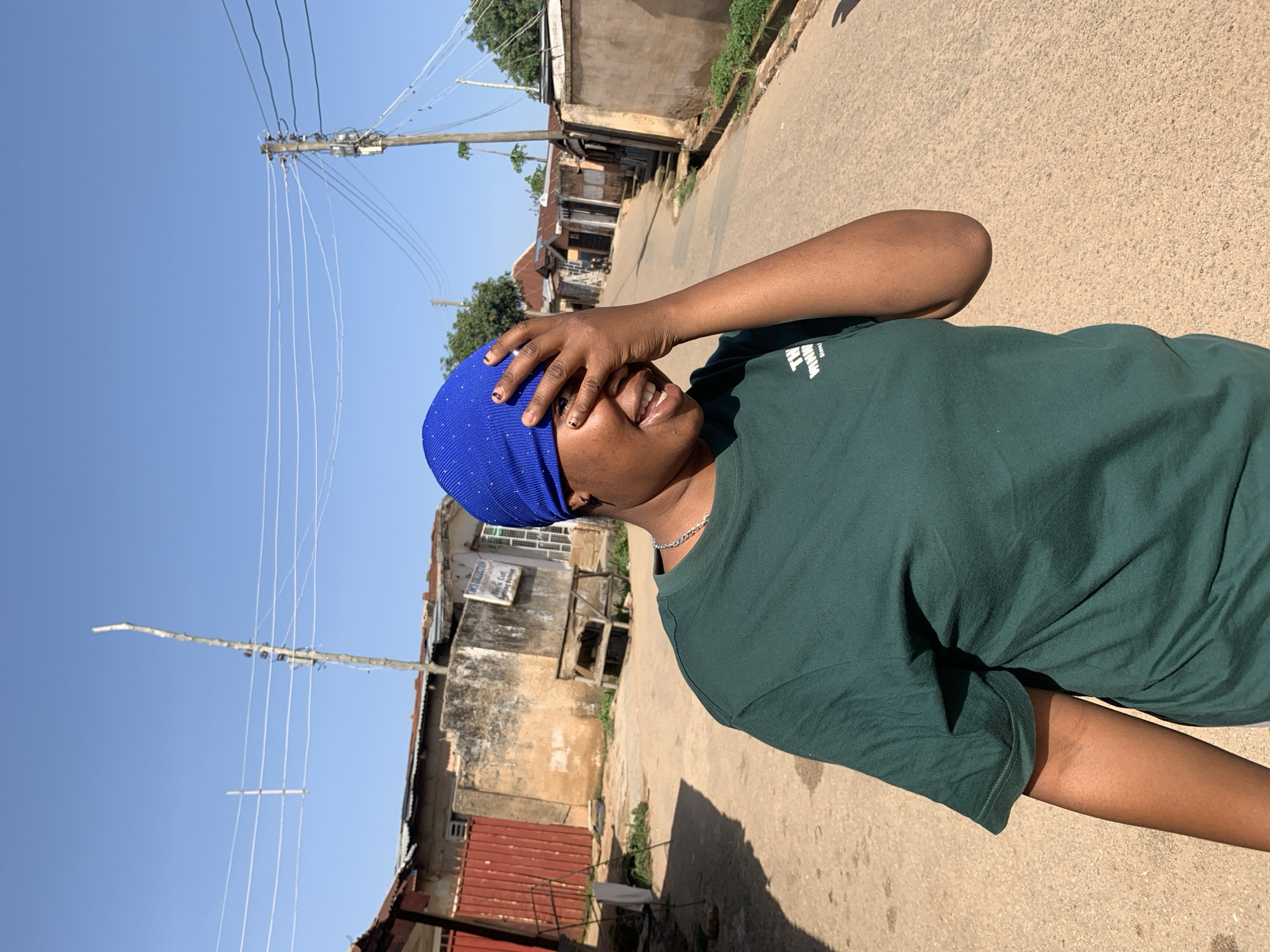 In an outdoor, sunlit street scene in the city of Jos, Nigeria, a young woman with dark skin stands in the foreground. She is wearing a dark green t-shirt and a bright blue head covering adorned with small white dots. Her right hand is covering her face, with her fingers partially obscuring her eyes and forehead. Her mouth is open, and she appears to be laughing or smiling. Her left arm is bent at the elbow and rests at her side. Behind her, the street is paved with asphalt and appears somewhat worn. On either side of the street are buildings with varying architectural styles, many appearing to be made of concrete or plaster, with corrugated metal roofs. Some of the buildings show signs of wear and age. Electrical wires are visible strung across utility poles in the background, typical of urban infrastructure. The sky is a clear, bright blue, indicating a sunny day. The overall impression is that of a candid moment captured on a street in a bustling, possibly residential or mixed-use area of Jos.