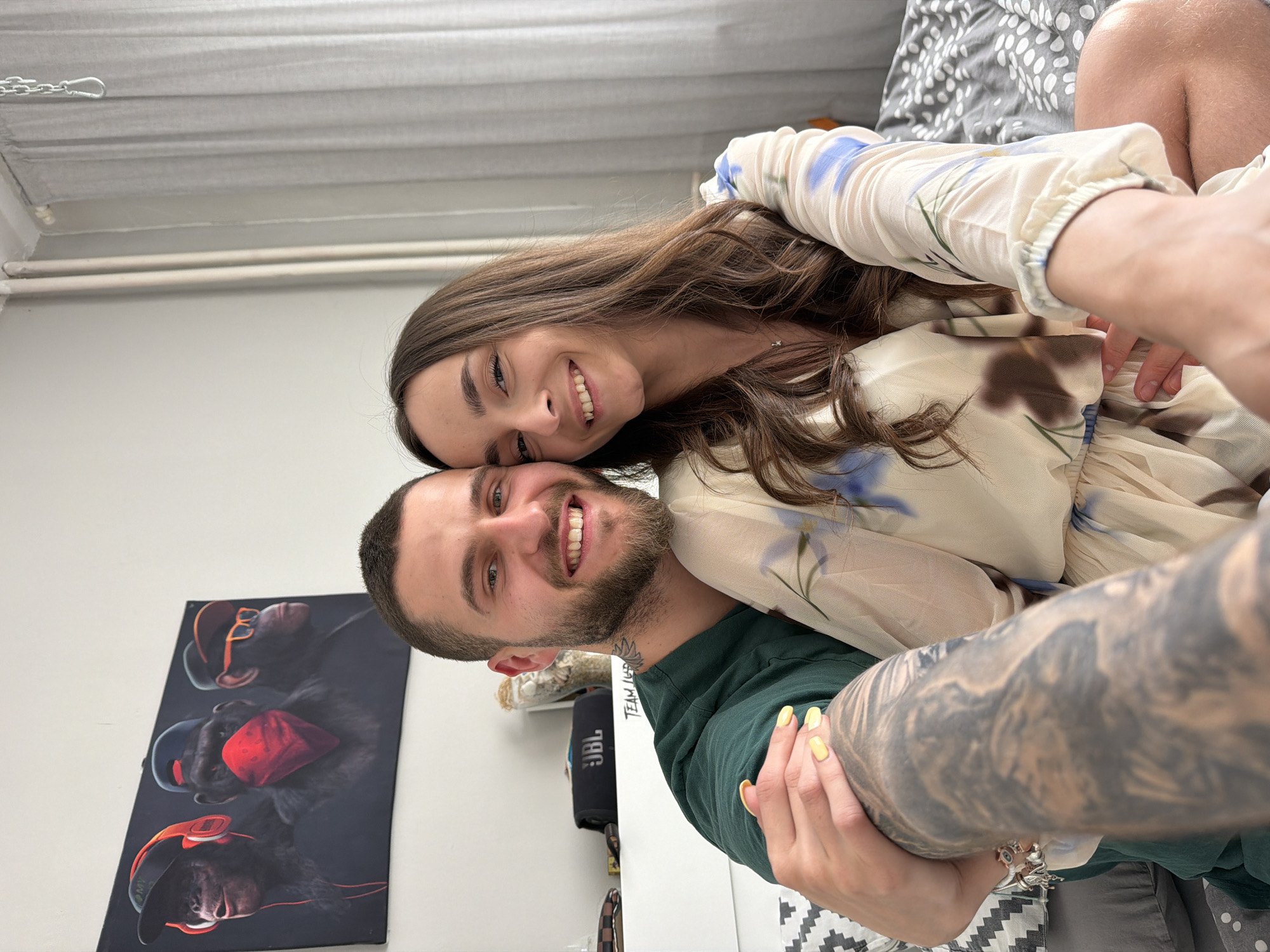 A man and woman are indoors, posing for a selfie. The man, with a short beard and green shirt, has an arm with a full sleeve tattoo wrapped around the woman. The woman, with long brown hair and a floral-patterned dress, is smiling and leaning her head against the man's. Behind them is a white wall with a painting of three monkeys wearing hats and masks. A black JBL speaker is visible near the man. The image's location within Sofia, Bulgaria, cannot be determined from the given visual information.