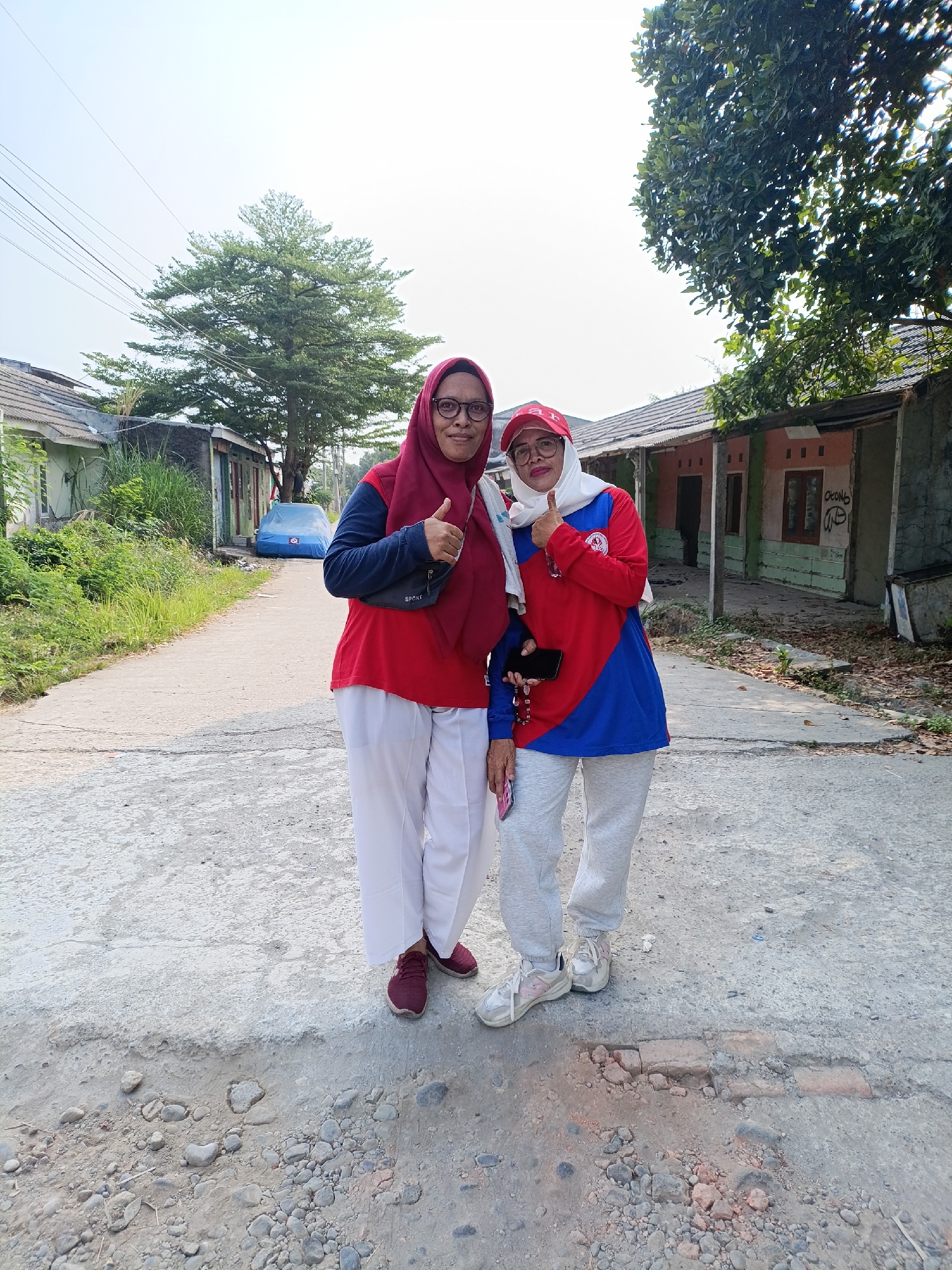 Two adult women are standing on a paved road in what appears to be a rural area with some houses and vegetation. The woman on the left wears a red hijab, glasses, a red long-sleeved shirt, and white pants. She has her left thumb raised in a "thumbs up" gesture. The woman on the right wears a red and blue long-sleeved shirt, a white hijab, and light gray pants. She also has her right thumb raised in a "thumbs up" gesture and is wearing glasses. Both women are wearing athletic shoes. In the background, there are several single-story houses, some with corrugated metal roofs, and lush green trees. A blue car is parked on the side of the road. The road surface is asphalt with visible cracks and gravel. The overall setting suggests a casual outdoor activity in a suburban or village environment.