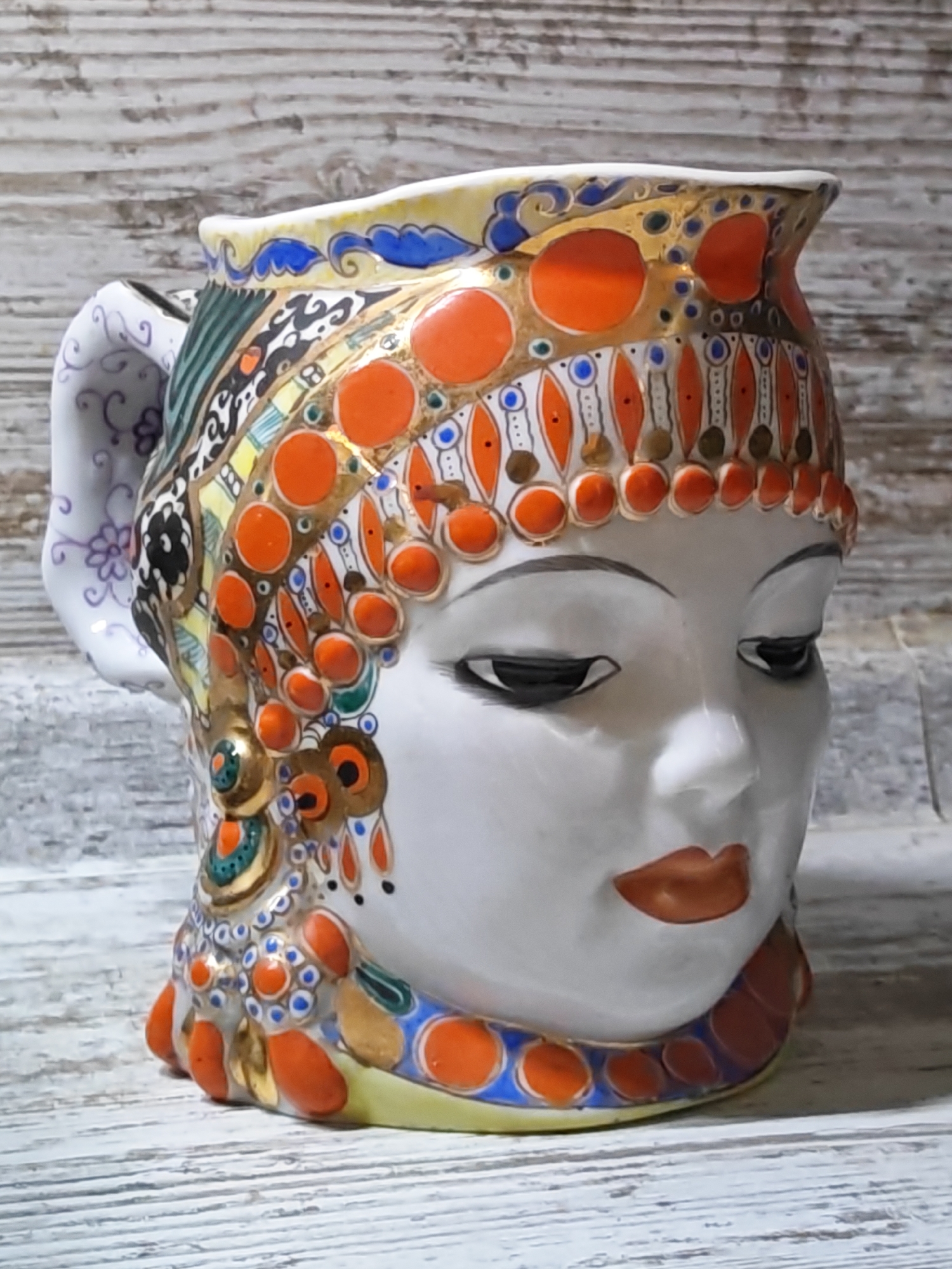 A ceramic pitcher shaped like a woman's head is displayed on a weathered wooden surface. The pitcher is adorned with intricate, colorful patterns featuring orange, blue, gold, and green hues. The face on the pitcher has closed eyes and painted lips. The background consists of distressed wooden planks. There is no information in the image to suggest a location in Voronezh, Russia.
