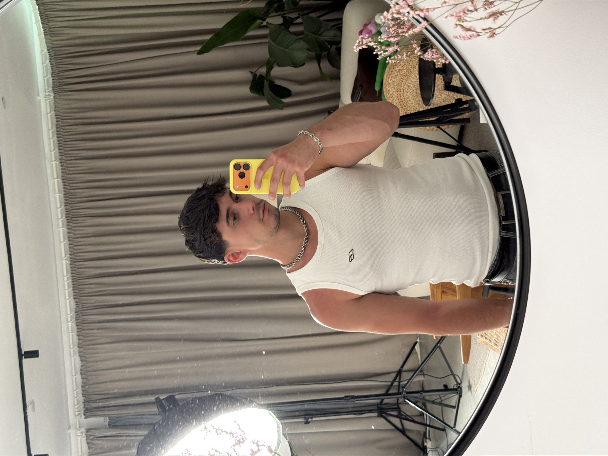 A young adult male in Sofia, Bulgaria, is depicted taking a mirror selfie indoors. He has short, dark, textured hair and is wearing a white ribbed tank top featuring a small black rectangular logo on the left chest. A silver-colored chain necklace adorns his neck, and a matching chain bracelet is on his right wrist. He holds a bright yellow smartphone with an orange camera module in his right hand, pointing it towards the mirror. The background, visible as a reflection, reveals light brown pleated curtains, a large green leafy plant, and various furnishings including a light-colored sofa, a dark round side table, a woven basket, and a segment of a wooden chair. A prominent octagonal softbox light on a stand is reflected on the left side, indicative of a photography setup. Black track lighting fixtures are visible on the white ceiling above the curtains. The mirror's thin black frame is visible at the bottom, and a vase containing pink dried flowers is partially reflected on the lower right.