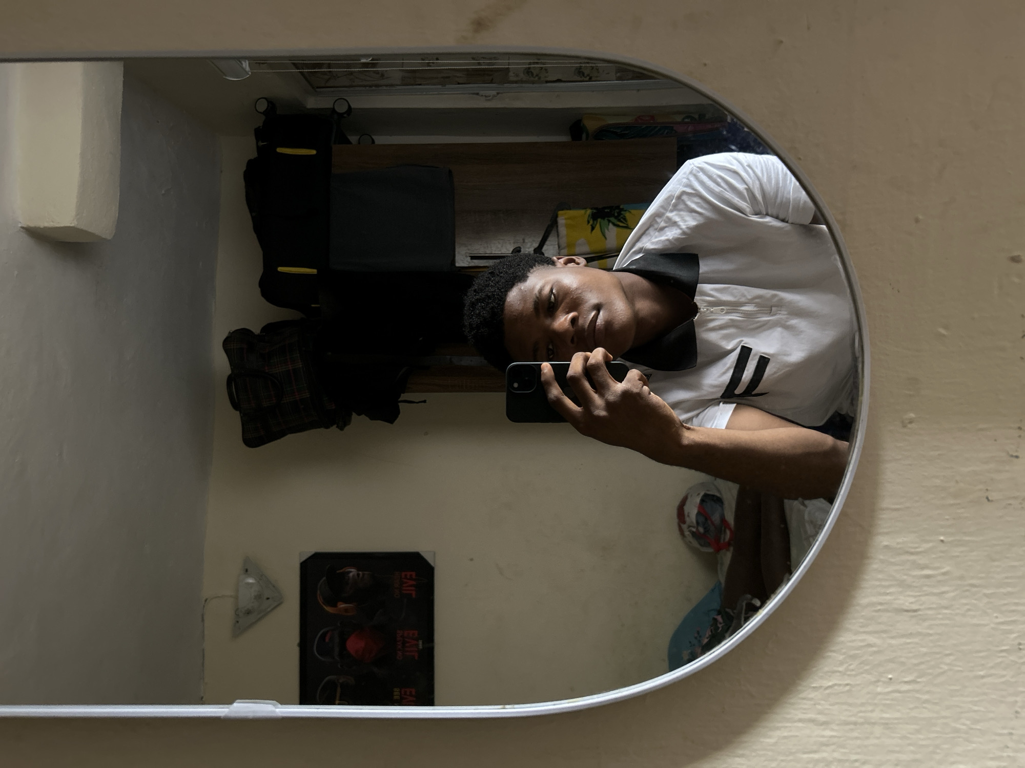 A young male is captured in a mirror selfie within an indoor setting in Ihiagwa, Nigeria. He is positioned centrally, holding a black smartphone with his right hand to photograph his reflection. He wears a white polo shirt featuring a black collar and two horizontal black stripes on the left chest. His hair is dark and closely cropped. The mirror is oval-shaped with a white frame.
Reflected in the mirror, the background reveals a light-colored, textured wall. On the left, a framed poster displays three stylized figures: one with a red face mask, another with orange headphones, and the third wearing a cap. Reversed text, "33E" and "EVIT," is visible on the poster. Above the poster, a white triangular ceiling light fixture is installed.
To the right of the poster, various items are visible on what appears to be a shelf or top of a wardrobe: a plaid patterned bag on the left and several black bags, at least one with yellow vertical accents. A wooden wardrobe unit is partially visible on the right side of the reflection, with a dark garment hanging from it. Below the poster, on a surface resembling a bed, a blue flat object and a white object with red accents (possibly a helmet) are present. A vertical blind or curtain system is partially visible on the far right. The lighting in the room appears ambient and somewhat subdued.