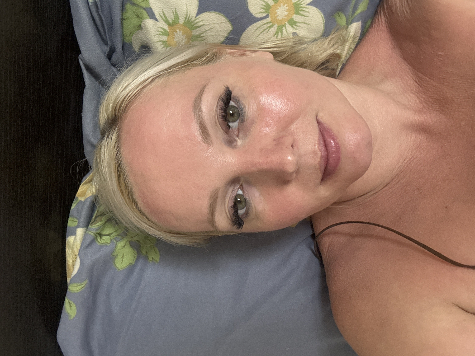 A medium close-up selfie shows a fair-skinned blonde woman with green eyes and light makeup lying on a floral-patterned pillow. Her head is angled slightly to her right. The pillow is blue with green leaves and white and yellow flowers. The upper portion of the image shows a dark, textured surface. The woman is wearing a thin spaghetti strap. Her skin appears to have a pinkish hue, possibly from a tan or sunburn. She has on false eyelashes and lip gloss. The context does not provide enough information to determine the location in Komsomolsk-on-Amur, Russia.