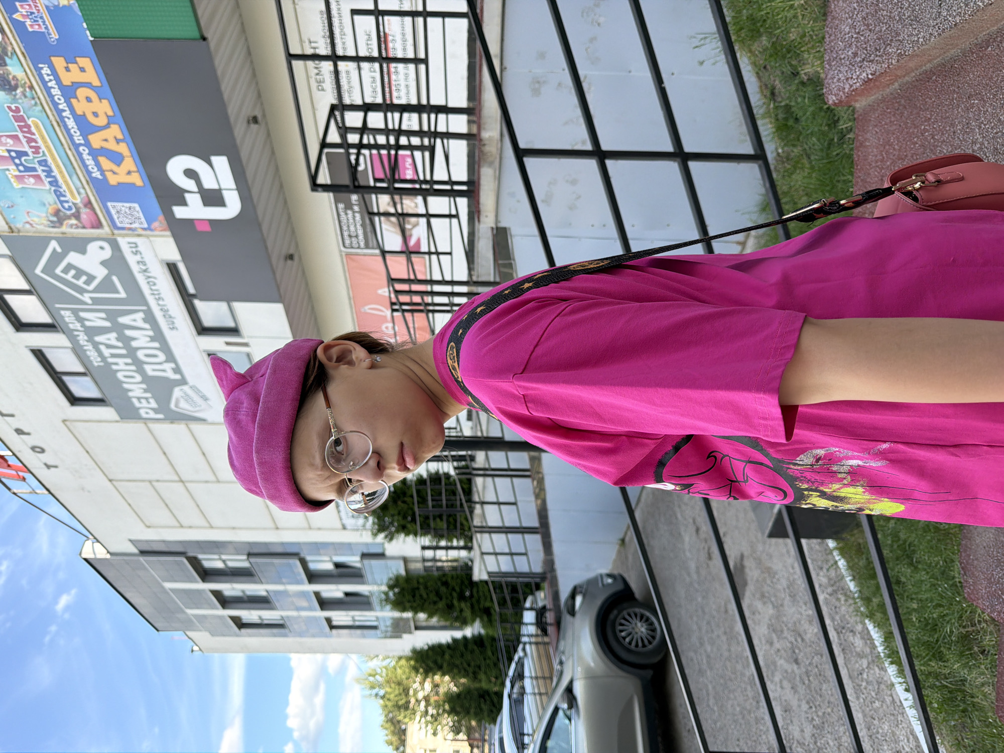 A young individual is captured outdoors, seen from the mid-torso upward, turning their head to look over their left shoulder directly towards the camera with a neutral to slightly stern expression. They wear a bright fuchsia beanie featuring a small, pointed projection on the left side, and round-rimmed spectacles with thin, light-colored frames. Their attire consists of an oversized, bright fuchsia t-shirt with a partially visible graphic design on the left side, displaying abstract yellow and lime green splatter elements over a darker outline. A dark strap, possibly embellished with golden stars, crosses their right shoulder, supporting a small, light peach-colored cross-body bag. A small, silver-toned stud earring is visible in their left earlobe.
The background features a multi-story commercial building under a clear blue sky with scattered white clouds. Prominent signage on the building includes Russian text: a partially visible "ТОРГ" (likely part of 'ТОРГОВЫЙ ЦЕНТР' - Shopping Center) at the top, "ТОВАРЫ ДЛЯ ДОМА" (GOODS FOR HOME) with a paintbrush and house logo, and the website "superstroyka.su". Other signs denote "СТРАНА ЧУДЕС" (LAND OF WONDERS) above a colorful graphic, "ДОБРО ПОЖАЛОВАТЬ!" (WELCOME!) above large yellow text "КАФЕ" (CAFE), and a distinct "t2" logo in white with a pink '+' symbol on a black background. Further signs indicate "РЕМОНТ" (REPAIR) services for "телефонов" (phones) and "ноутбуков" (laptops), along with "Часы работы:" (Opening hours:).
To the left of the individual, a silver-grey SUV is parked. Behind the vehicle and further into the distance, several dark green coniferous trees are visible. The immediate foreground and mid-ground are characterized by dark metal railings, suggesting a barrier or part of a stairway/ramp. The ground comprises concrete pavement, patches of green grass, and a reddish-brown textured surface in the bottom right corner. The scene is situated in a commercial district within the city of Chaikovsky, Russia.
