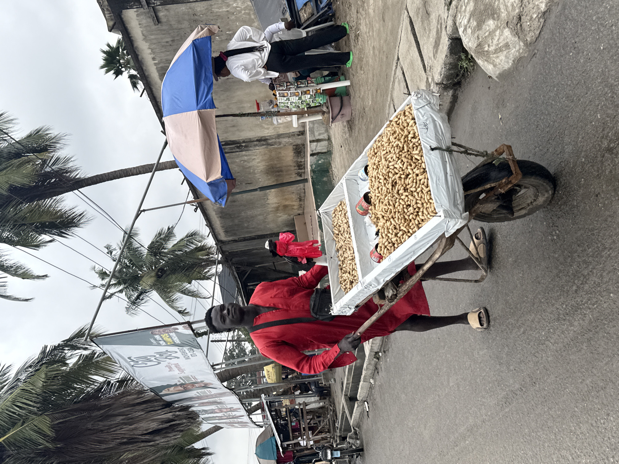 In Ado, Nigeria, a man in a red tunic pushes a wheelbarrow filled with peanuts down a paved street. The wheelbarrow has a single wheel and is supported by two wooden handles. The man is wearing simple sandals. To the right of the man, a stall is set up with an umbrella providing shade. A woman in a white shirt and black pants stands at the stall, and another person is partially visible behind her. Various goods are displayed on shelves at the stall, and a pink bag is visible in the background near a stall to the left. Palm trees are present, and the sky is overcast. The street has a concrete curb and a drainage channel. Several motorcycles are parked on the left side of the image near some stalls. A banner advertising "City Boy" and "Movement" is visible in the background, suspended between poles.