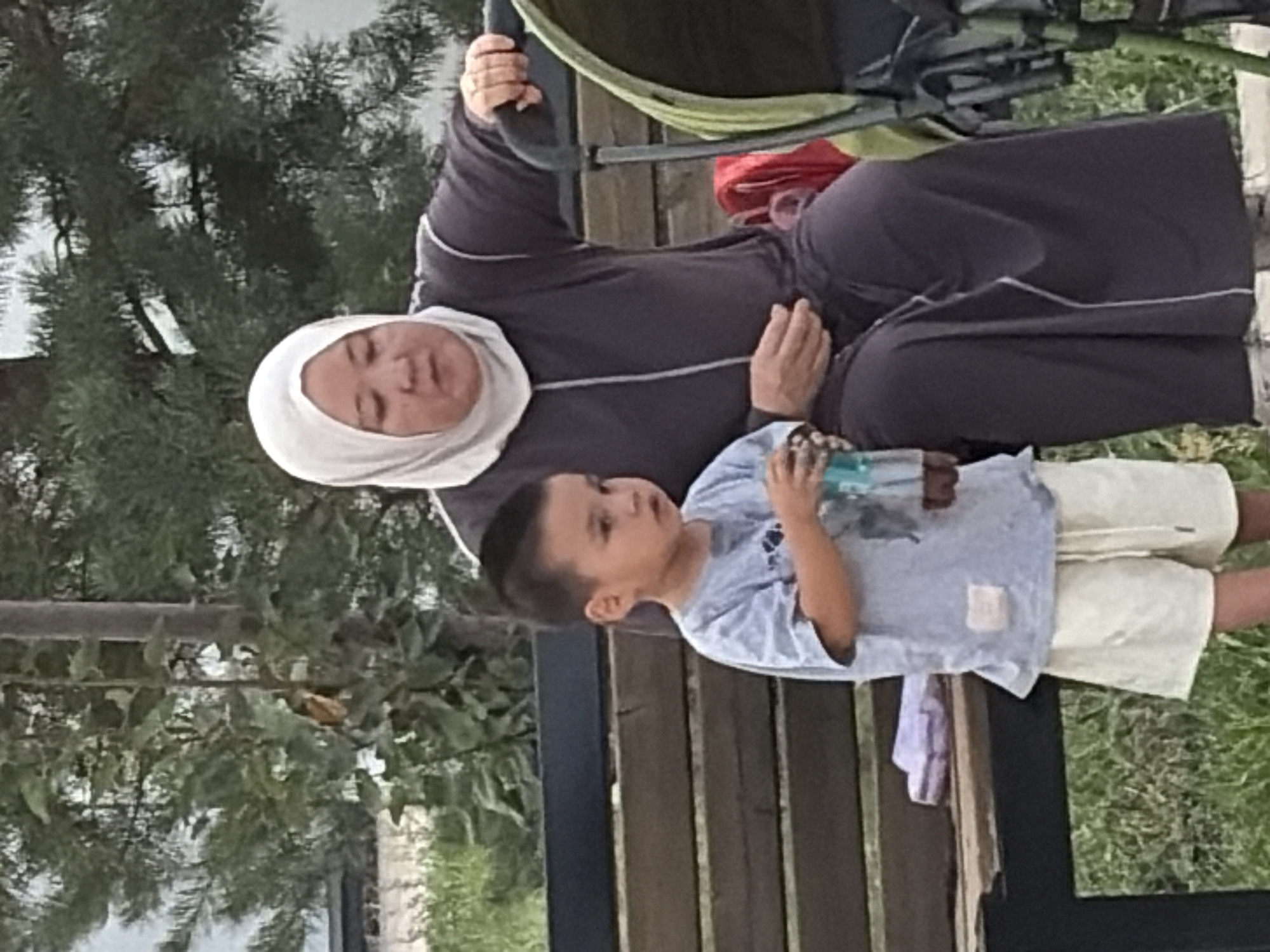 A medium shot captures a young boy and a woman seated on a wooden park bench. The boy, on the left, is wearing a light blue t-shirt and off-white shorts. He is holding a bottle of water in his right hand and looking to his left. The woman, on the right, is wearing a dark grey, long-sleeved garment and a white hijab. She is looking down and to her left. Her right hand rests on the handle of a stroller, which is partially visible on the right side of the frame. A red bag is tucked behind her. The background consists of greenery and trees, suggesting an outdoor park setting. The lighting indicates it is daytime.