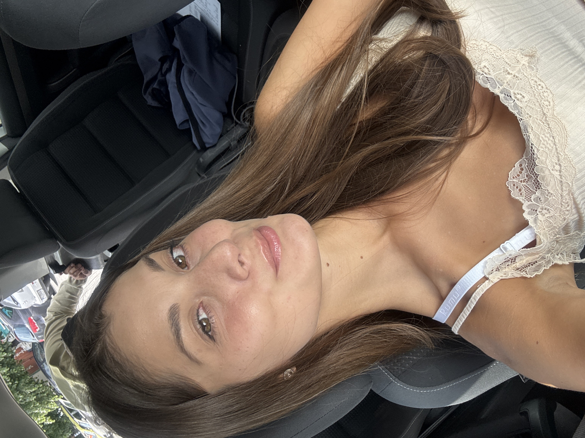 A young woman with brown hair and light skin is lying in the passenger seat of a car. She is wearing a light-colored top with lace detailing and a visible bra strap. The car seat is black, and there is a blue garment in the background. Outside the car window, there is a partial view of a street scene with cars and trees, suggesting an urban environment. It is not possible to ascertain if the photo was taken in Burgas, Bulgaria.