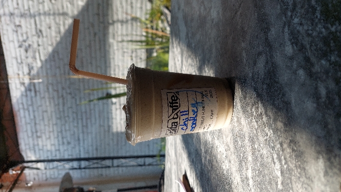 The image depicts a close-up, static view of a plastic beverage cup in an outdoor or semi-outdoor commercial setting in Tlaxcala City, Mexico. The central object is a clear plastic cup containing a light brown, frothy, blended beverage, with a light brown, bent plastic straw inserted. A white rectangular label affixed to the cup displays "Vita Caffe" at the top and the handwritten blue text "chill monkey". Below this, partially visible text includes "SABANA HUNGARINA TLX" and "CAFE", specifically identifying the location as a Vita Caffe branch on Sabana Húngara street in Tlaxcala City. The cup rests on a dark grey, textured surface, likely a tabletop, with strong, directional sunlight creating sharp shadow patterns. In the background, a light-colored brick wall is visible, with minimal green foliage behind the cup. A dark metal structural element is partially visible in the upper left, suggesting an awning or canopy overhead. No individuals, actions, or direct interactions are present in the scene.