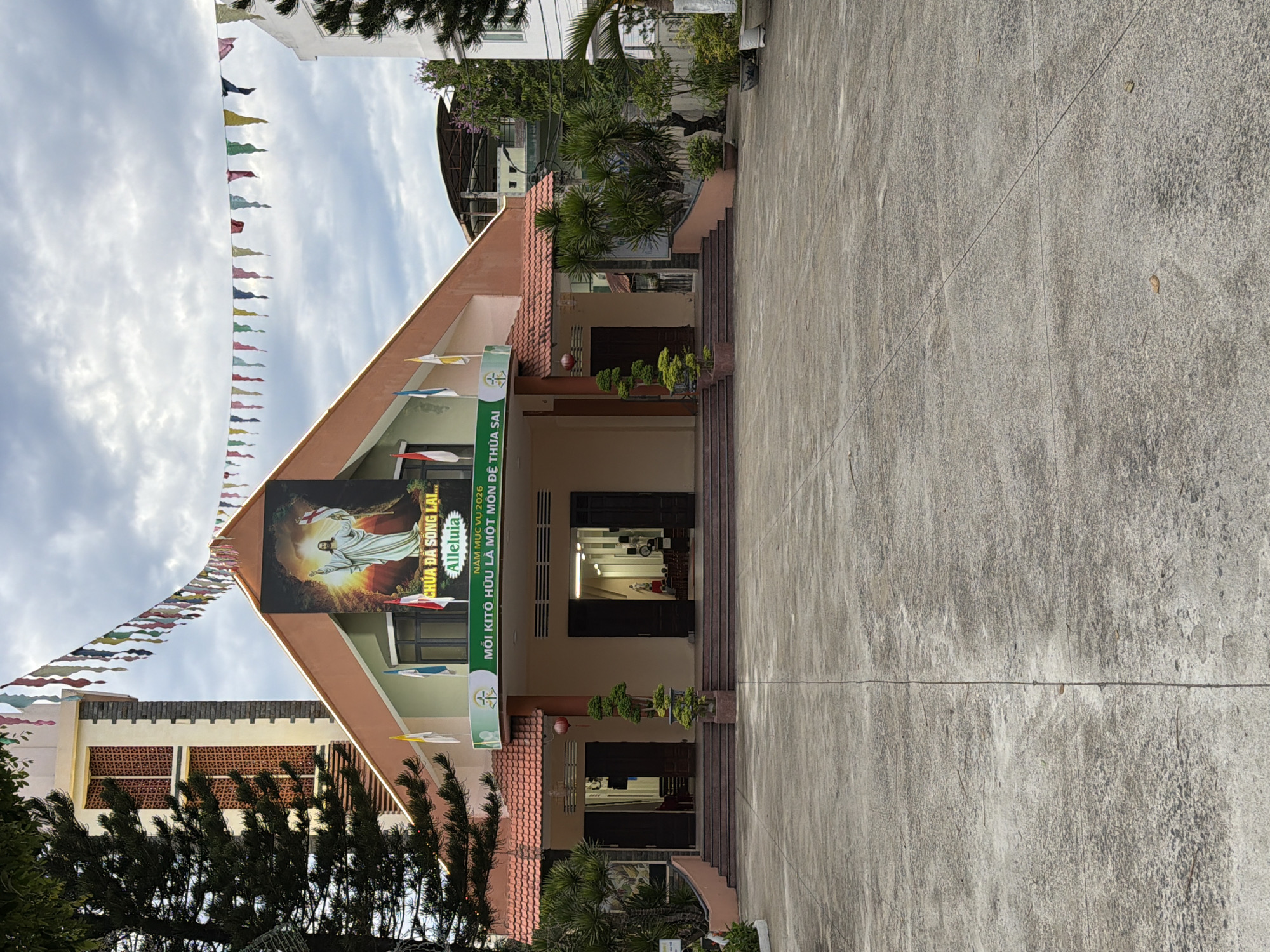 The image shows the front of a church in Đà Nẵng, Vietnam, identifiable by the text "CHÚA ĐÃ SÔNG LAI..." and "NĂM MỤC VỤ 2026." A large image of Jesus Christ is displayed on the building's facade. The entrance is flanked by steps and potted plants. A series of small, colorful triangular flags are strung across the front of the church, creating a festive appearance. The foreground is dominated by a concrete area leading up to the church.