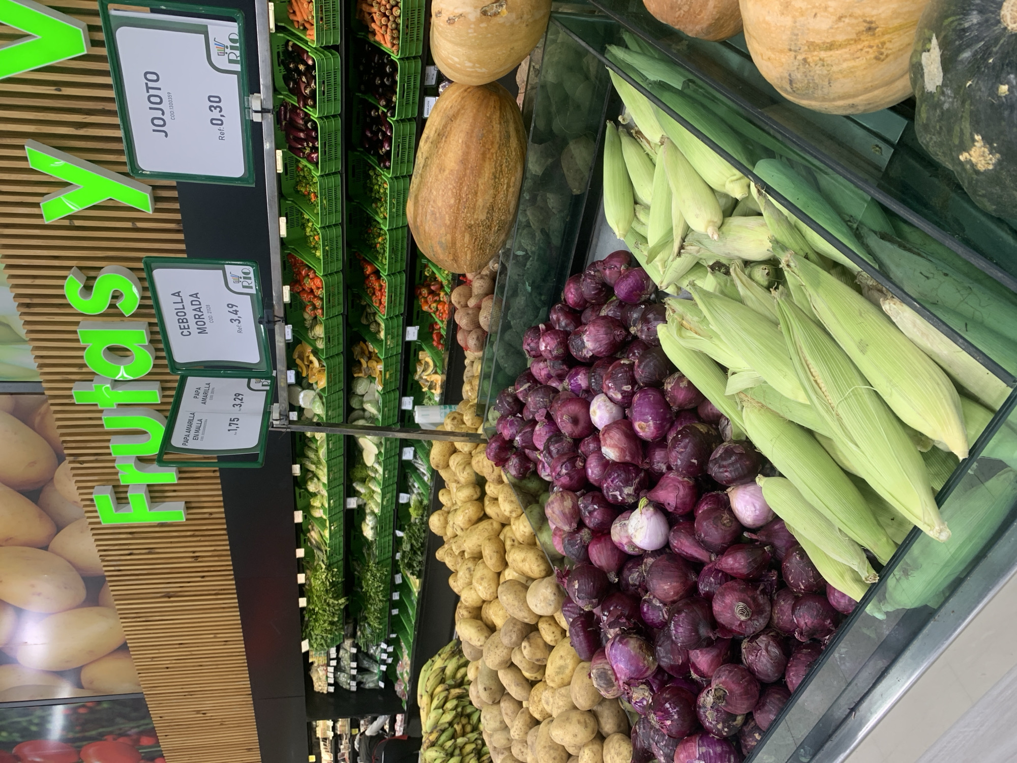 This image captures a vibrant and well-stocked produce section of a grocery store in Juan Griego, Venezuela. The scene is brightly lit, showcasing an abundant display of fresh fruits and vegetables.
In the foreground and mid-ground, large piles of purple onions are prominently featured, alongside numerous ears of corn still in their green husks. Next to the onions, a generous mound of yellow potatoes is visible. Above these items, on a higher shelf, several large gourds or pumpkins of varying sizes and colors are displayed. The background reveals multi-tiered green shelves packed with a diverse array of other vegetables in green plastic crates, including what appear to be carrots, eggplants, bell peppers, and various leafy greens, suggesting a wide selection available to customers.
No people are visible in the frame, emphasizing the static display of merchandise. The store appears organized and well-maintained, with produce neatly arranged.
Several price tags and signs are visible, written in Spanish, serving as location cues. A large, bright green sign on the left partially spells out "Frutas y" (Fruits and). Individual price tags identify products:
- "JOJOTO" (corn) with "COD.1300359" priced at "Ref: 0,30".
- "CEBOLLA MORADA" (purple onion) with "COD. 21128" priced at "Ref: 3,49".
- "PAPA AMARILLA" (yellow potato) with "EN MALLA" and "COD 2059", showing prices "1,75 3,29", with 3,29 being the more prominent value.
Each of these price tags also features a "Rio" logo. The wooden slat wall decor behind the "Frutas y" sign adds to the aesthetic of the store.