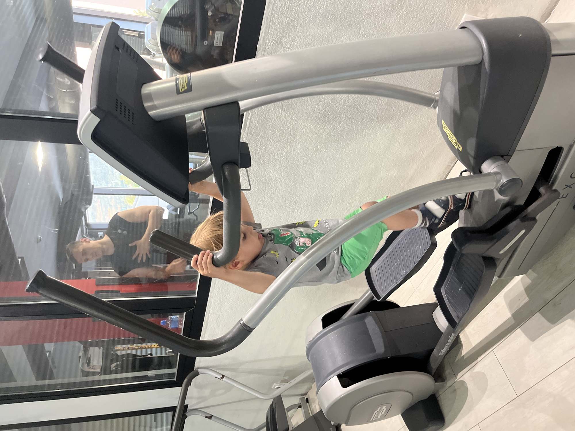 A young child is hanging from the handles of a Technogym elliptical machine in what appears to be a home gym or fitness center in Vranovići, Montenegro. The child is wearing a gray t-shirt with a dinosaur design and green shorts. The elliptical machine is gray and black and has a monitor attached. A man can be seen reflected in a glass window in the background, facing left while wearing a dark top. The surrounding environment includes a white wall, a window with a red and white wall visible outside, and a tiled floor. The image is rotated counter-clockwise.
