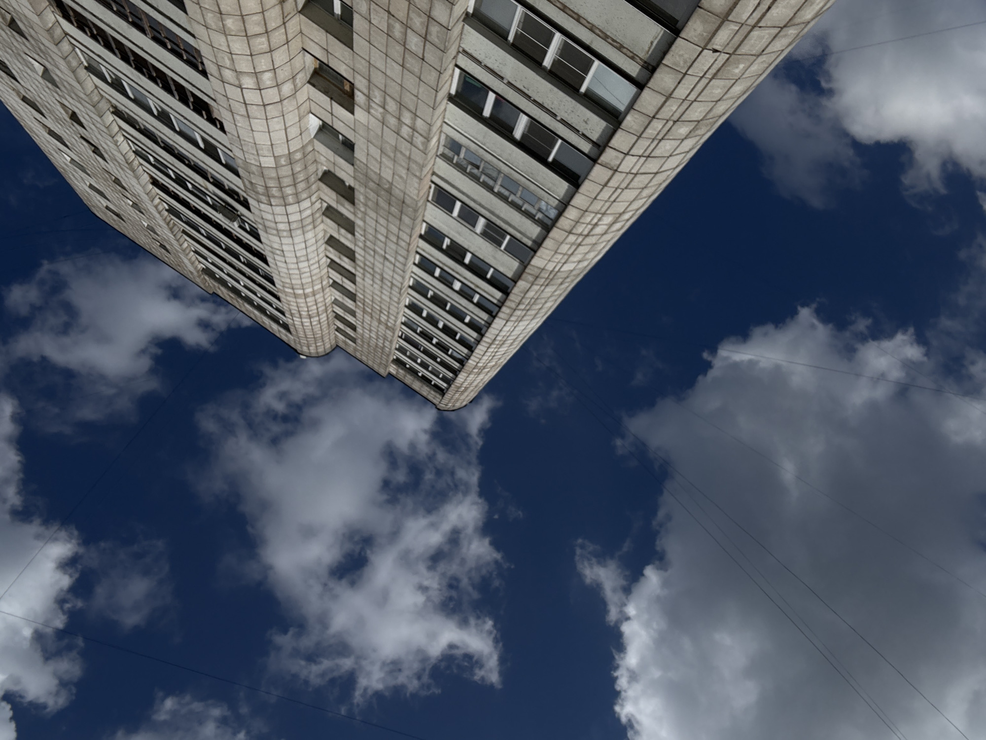 The image shows a low-angle shot of a tall, concrete apartment building against a dark blue sky with scattered white clouds. The building has a repetitive facade with rows of windows, and its structure appears to be curved or rounded on the side facing the camera. The perspective makes the building seem to stretch upwards and outwards, emphasizing its height and imposing presence against the sky. Thin wires are visible crisscrossing the sky in the foreground. The overall impression is one of a slightly overcast day, possibly in the afternoon, given the contrast between the dark sky and the bright clouds. There are no people visible, and the focus is on the architectural structure and the sky. The image was taken in Chaikovsky, Russia.