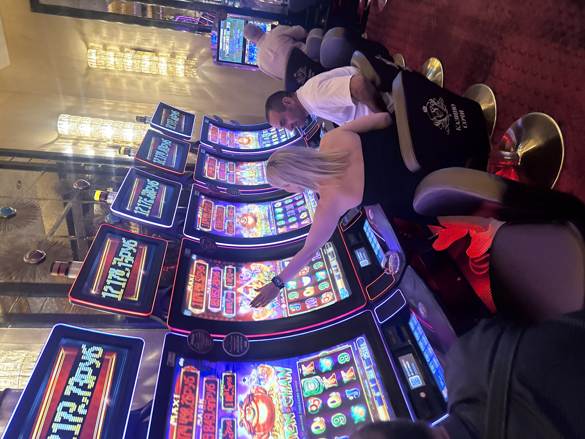 In the city of Estosadok, Russia, a group of individuals are seen at a casino, engaging with slot machines. The scene is dimly lit, with the neon glow of the machines illuminating the room.
In the foreground, a woman with blonde hair is facing away from the camera, her arm extended towards a slot machine, as if pressing a button or interacting with the screen. She is wearing a black strapless top. Beside her, a man with short dark hair and a white t-shirt is seated and looking towards the slot machines. His posture suggests he is also a participant in the gambling activity.
In the background, another man is seated at a slot machine, his back to the camera, wearing a light-colored shirt. Further back, more slot machines are visible, some displaying numbers and graphics in Russian. The machines have prominent screens showing monetary values and game information, with visible Russian text such as "МАЖИ" and "РУБ".
The environment appears to be a modern casino with plush seating, indicated by the presence of chairs with embroidered logos that read "КАЗИНО СОЧИ" (Casino Sochi). The floor is carpeted in red, and decorative wall lights contribute to the ambient lighting. The overall impression is one of a typical casino setting, focused on the activity of playing slot machines.
