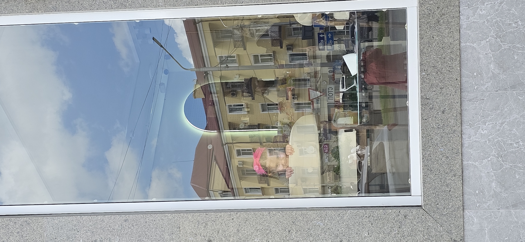 The image depicts a street scene reflected in a large window, likely a storefront, set within a grey, textured frame. The reflection shows a partly cloudy sky, a light pole, and a multi-story building with a peach-colored facade and a red roof. Other signs are reflected, including traffic signs, but these cannot be clearly read. A person, possibly a child, is visible partially obscured by the glass. Based on the presence of street signs in Russian and Cyrillic script, it is likely located in the city of Grozny, Russia.