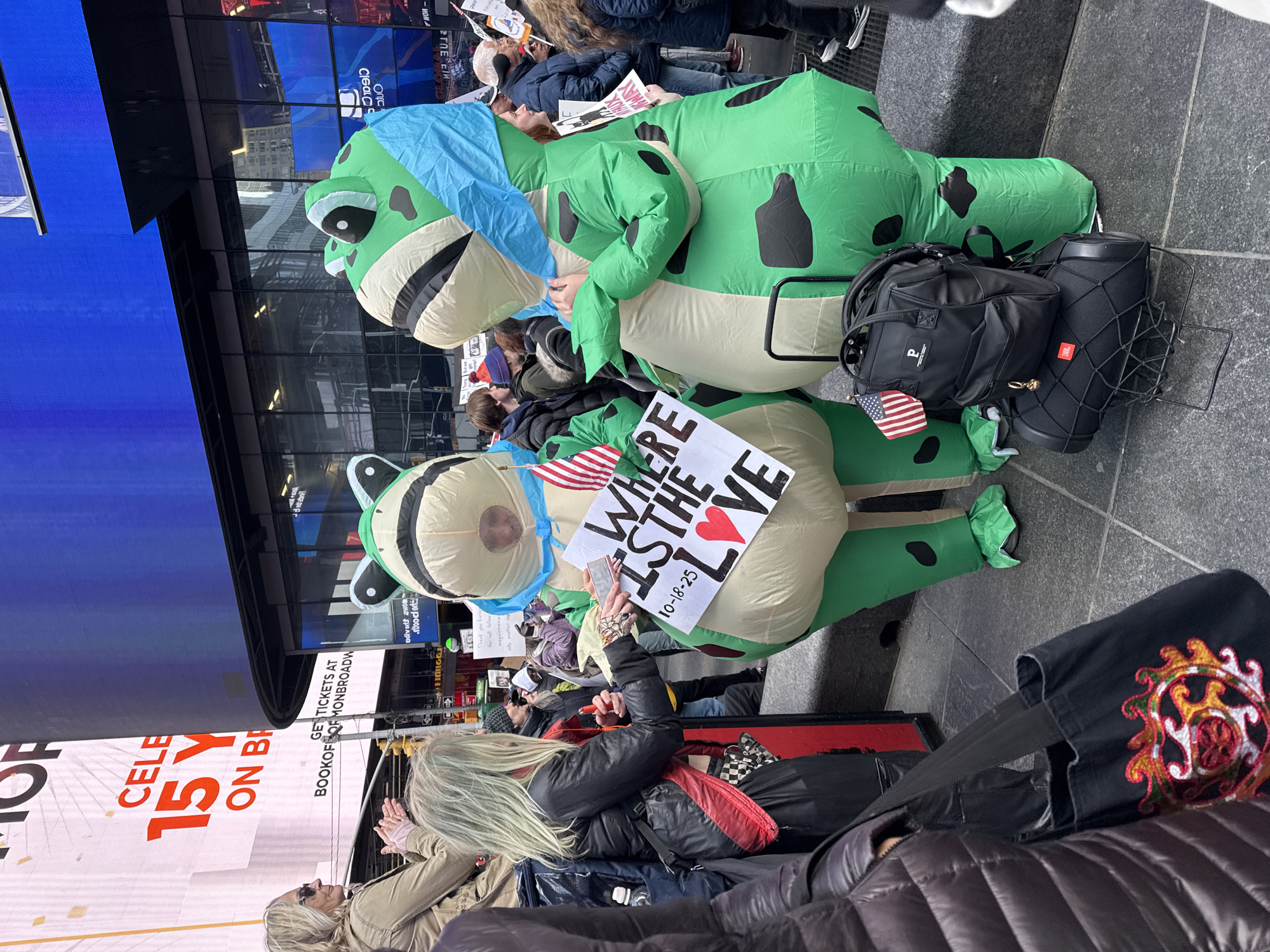 Here is a description of the image:
The image shows a crowded scene in what appears to be Times Square in New York City. Two people in inflatable green dinosaur costumes are the main subjects. One person holds a sign that reads "Where is the love" with a red heart and "10-18-23" written on it. A person with blond hair is taking a photo of the inflatable costumes and sign with their phone. There is a backpack and a JBL speaker next to one of the costumes. Billboards and advertisements are visible in the background, indicating the location. The general atmosphere seems to be that of a protest or demonstration.