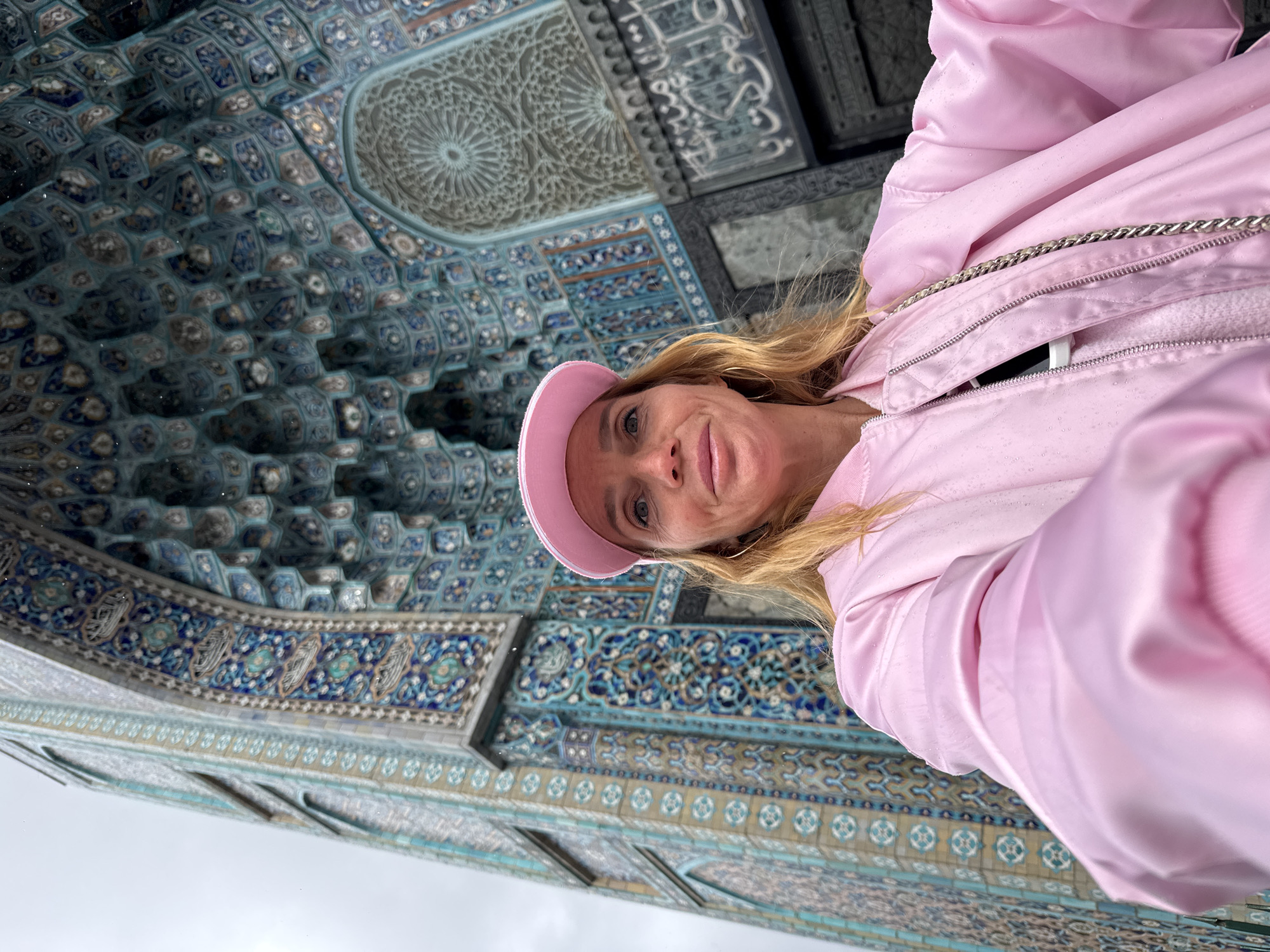 A woman wearing a pink jacket and matching pink hat is taking a selfie in front of the Saint Petersburg Mosque in Saint Petersburg, Russia. The mosque's detailed, blue-tiled architecture is prominently displayed in the background. The sky is overcast.