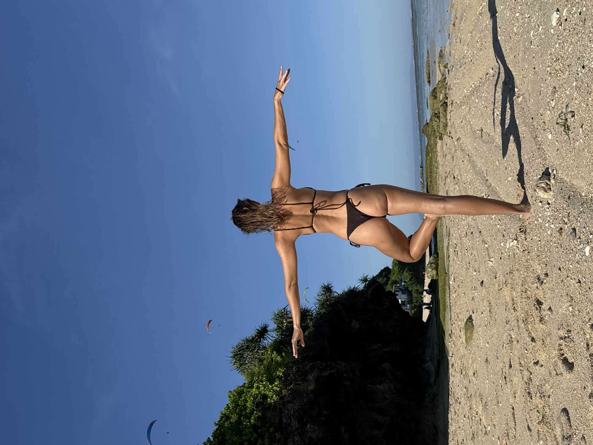 A woman in a dark bikini strikes a balancing pose on a beach in Kuta Selatan, Indonesia. She extends her right arm upward and her left arm downward for balance. The sky is clear and blue, with two paragliders visible. Behind her, a rocky, vegetated cliff meets the sandy beach. The beach extends to the horizon, where the ocean meets the sky. The woman's shadow is cast onto the beach. In the background, the architecture of a building is visible.