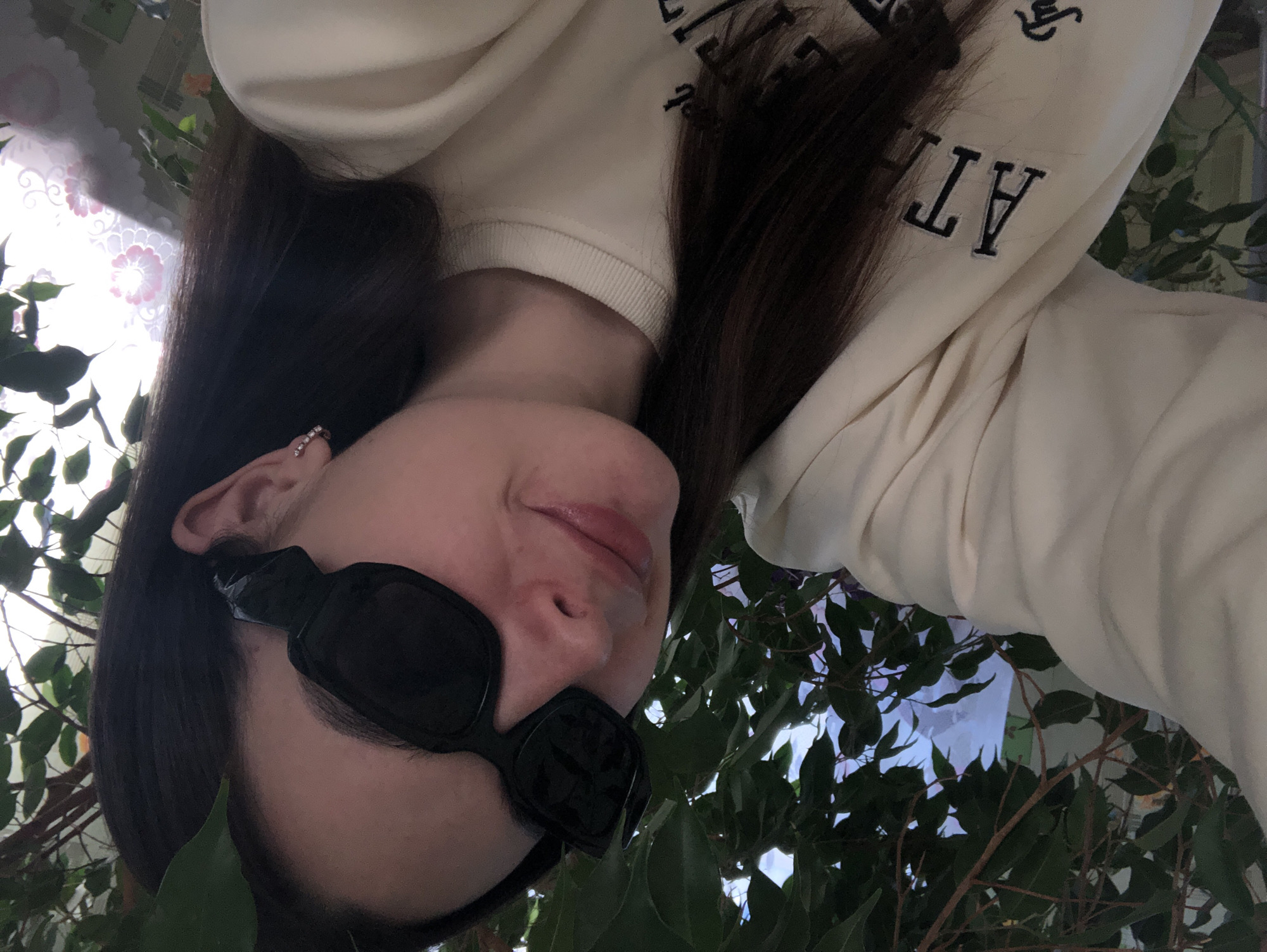 A woman with long, dark hair and fair skin is positioned at an odd angle, seemingly upside down, in front of a leafy tree or bush. She is wearing a light-colored sweatshirt with lettering on the sleeve, and black sunglasses. A small earring is visible in her left ear. The background includes glimpses of a window with a floral pattern. There is no visual information to confirm the location is Noviy, Russia.