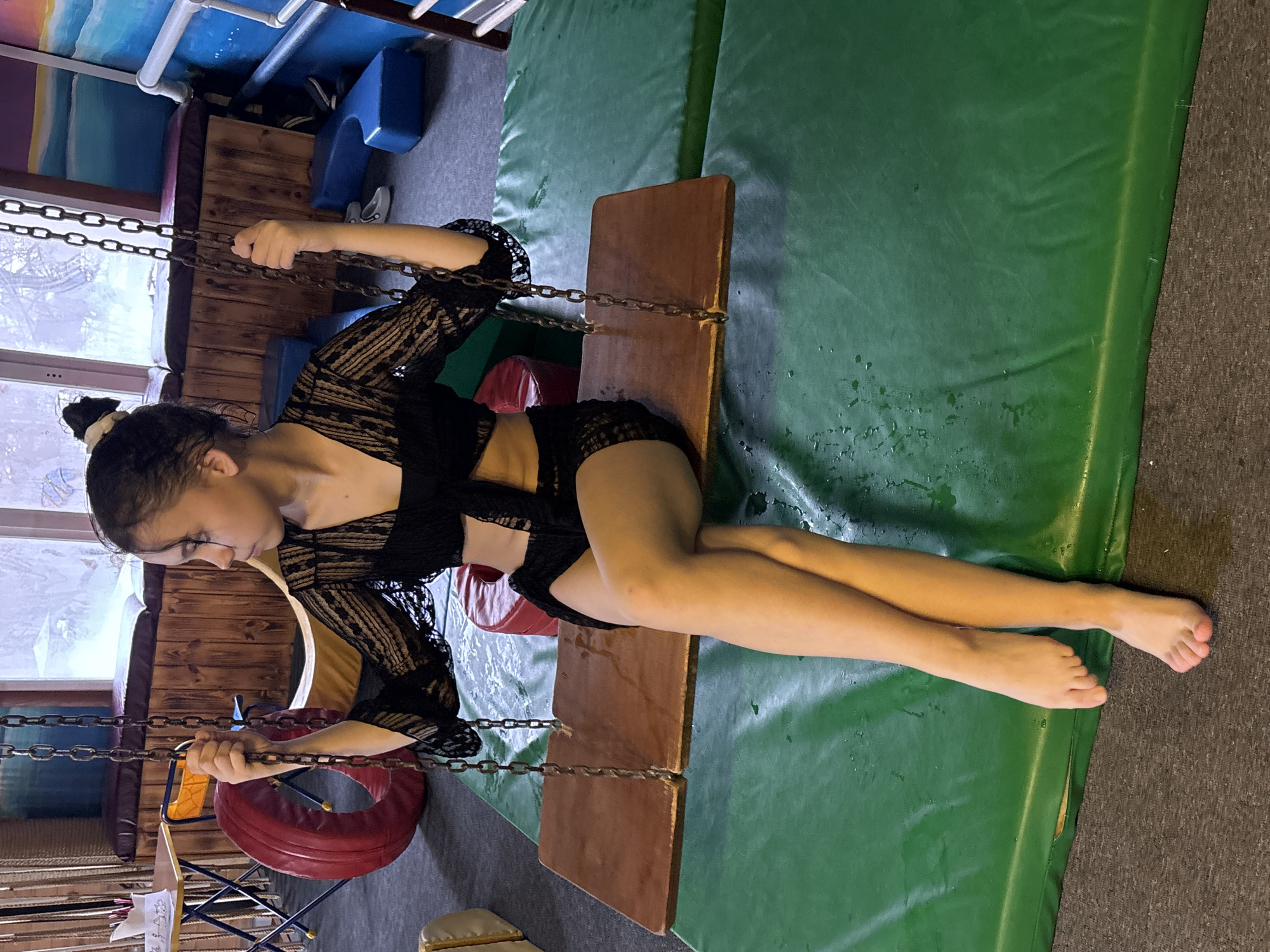 A girl sits on a swing with chains. She wears a dark, lacy top and dark shorts. The swing is set against a green padded floor or mat. The background includes gym equipment and window light. The location is Chaikovsky, Russia.