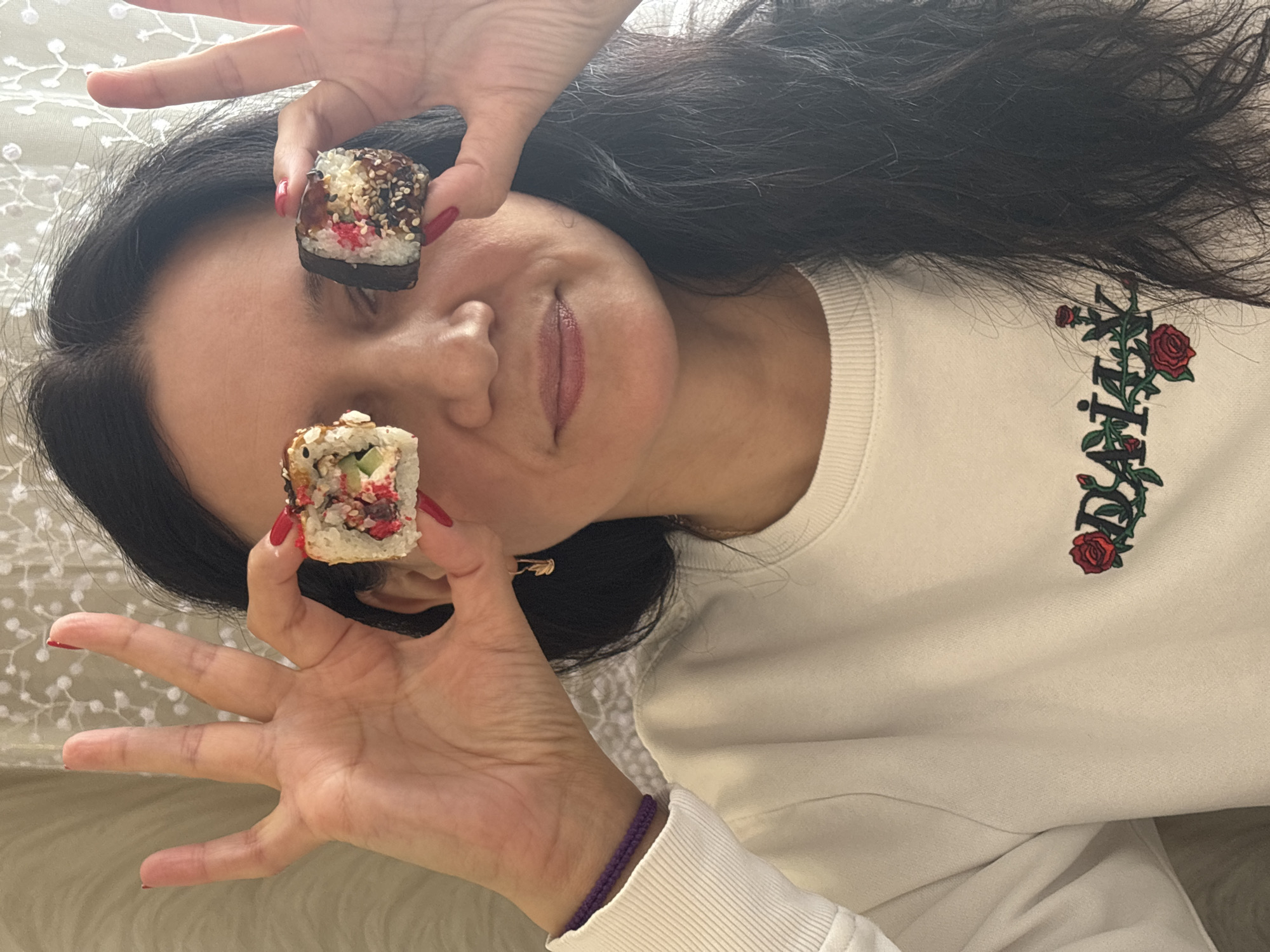 A woman with long, dark hair is holding two pieces of sushi in front of her eyes, using her hands to frame them. She is wearing a cream-colored sweatshirt with a floral design on the right side. Her fingernails are painted red. She is wearing a purple bracelet on her left wrist. The background appears to be a patterned fabric, possibly a bedspread. The photo was possibly taken indoors in Chaikovsky, Russia, though there is no definitive evidence of this within the image.