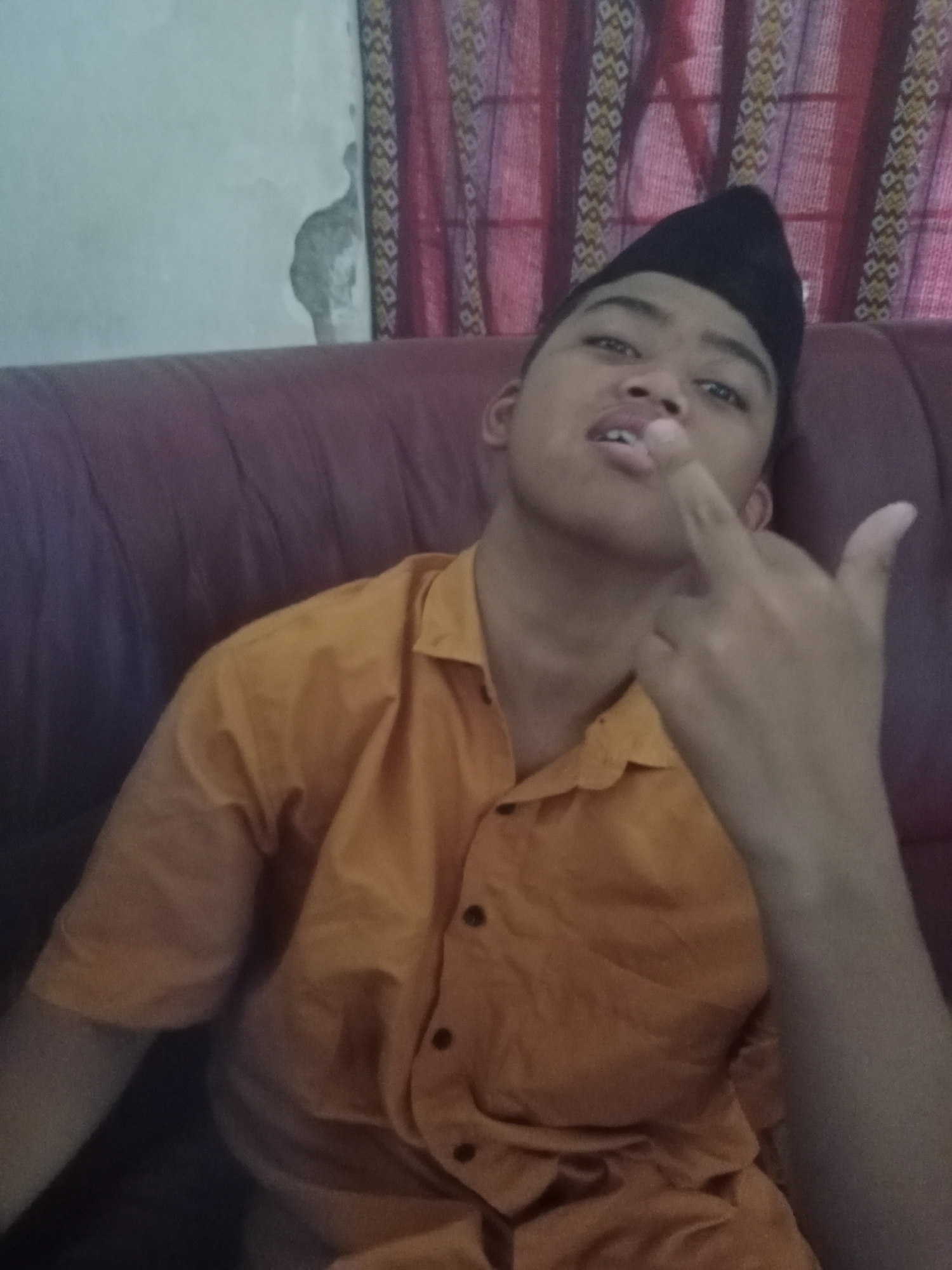 The image shows a young person sitting on a dark-colored couch, likely in a home environment. The person is wearing an orange collared shirt and a black cap. Their right hand is raised, with the middle finger extended. The background features a wall with peeling paint and red curtains with a patterned design. The lighting suggests an indoor setting, possibly during daytime. The overall scene is casual and informal. The location of Ringinrejo, Indonesia, is not discernible from the visual elements of the image itself.