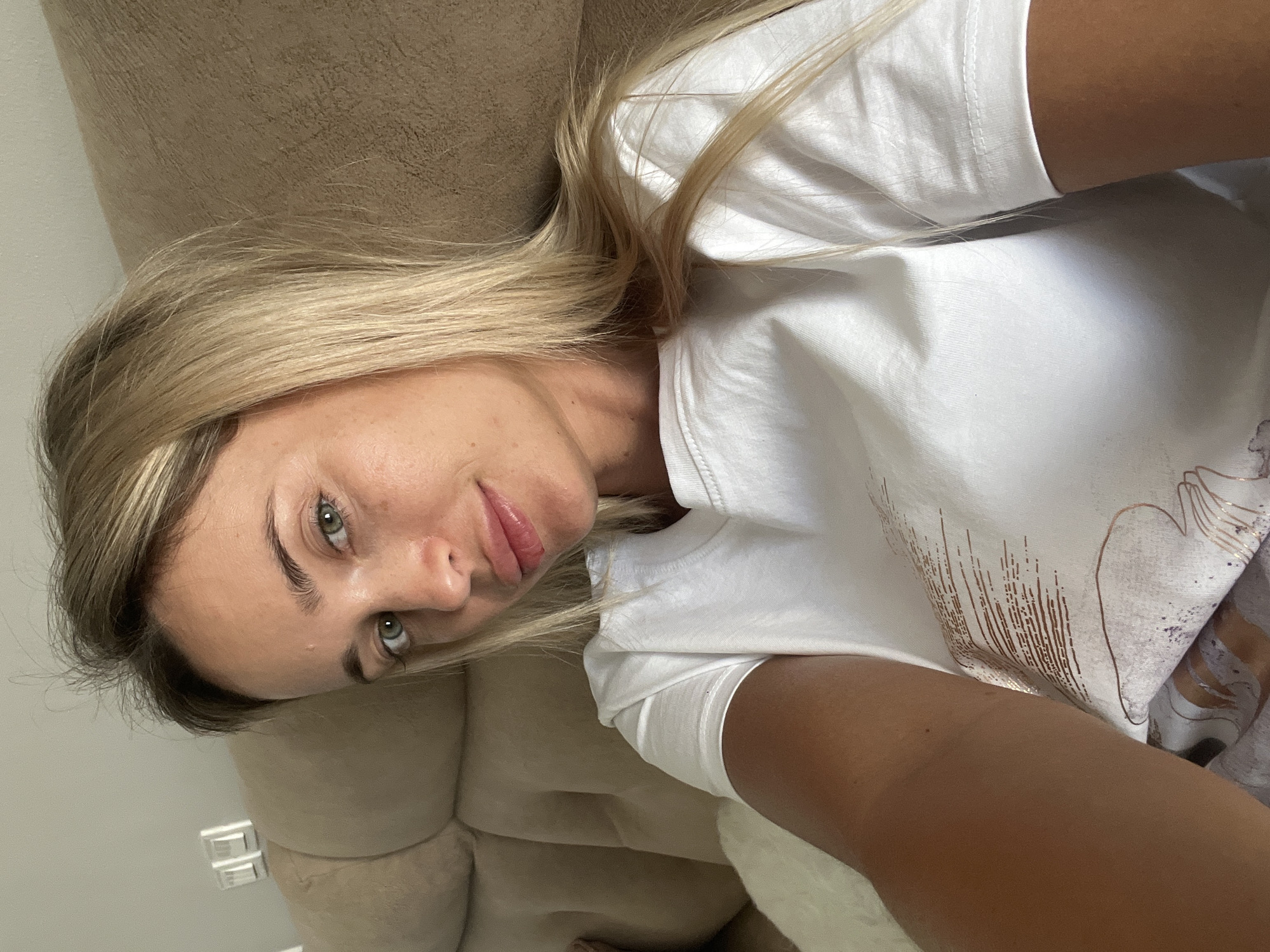 A young woman with blonde hair and green eyes, wearing a white t-shirt, is seated on a tan-colored sofa. The background appears to be an indoor setting, possibly a living room, with light-colored walls. One wall has what looks like electrical outlets. The woman is looking towards the camera, and her left arm is extended, holding what is likely a phone for a selfie. Her lips are slightly parted, and she has some makeup on. The image is a close-up shot, focusing on her face and upper body. There are no discernible landmarks or elements that would specifically place this scene in Sharm El Sheikh, Egypt.